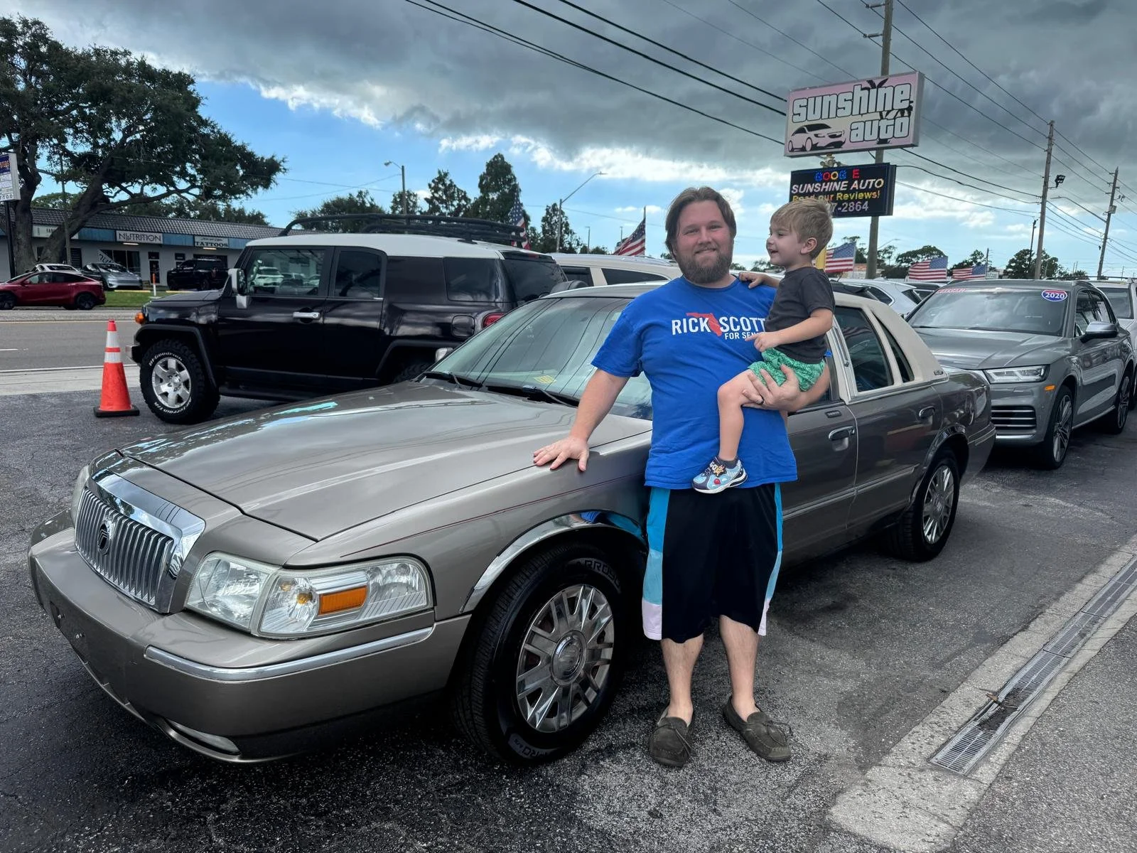 A man and a young boy standing in front of a beige luxury car at a car dealership. The man is wearing a blue "Rick Scott for Senate" t-shirt and black shorts, while the boy is dressed in a black t-shirt and green shorts. The man has his hand on the c