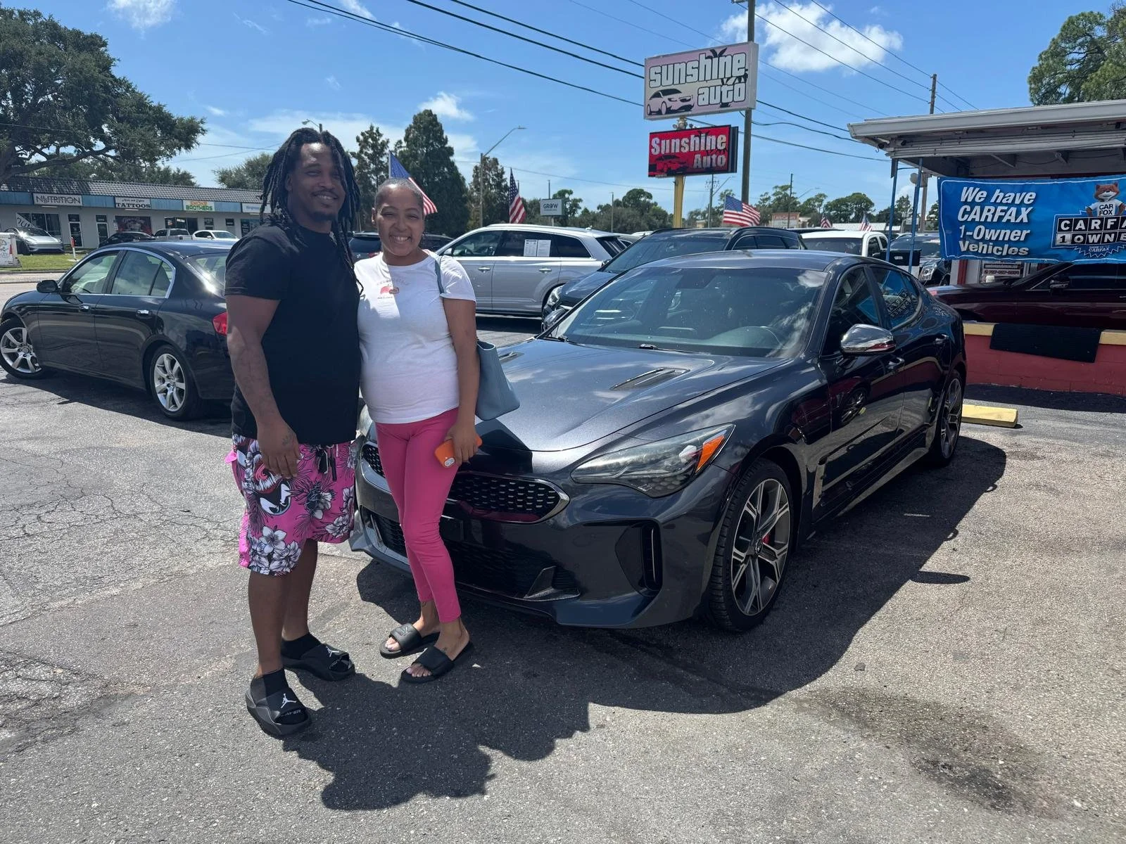 A man and a woman standing next to a dark gray sports car at a car dealership lot, with several other cars and dealership signage in the background.