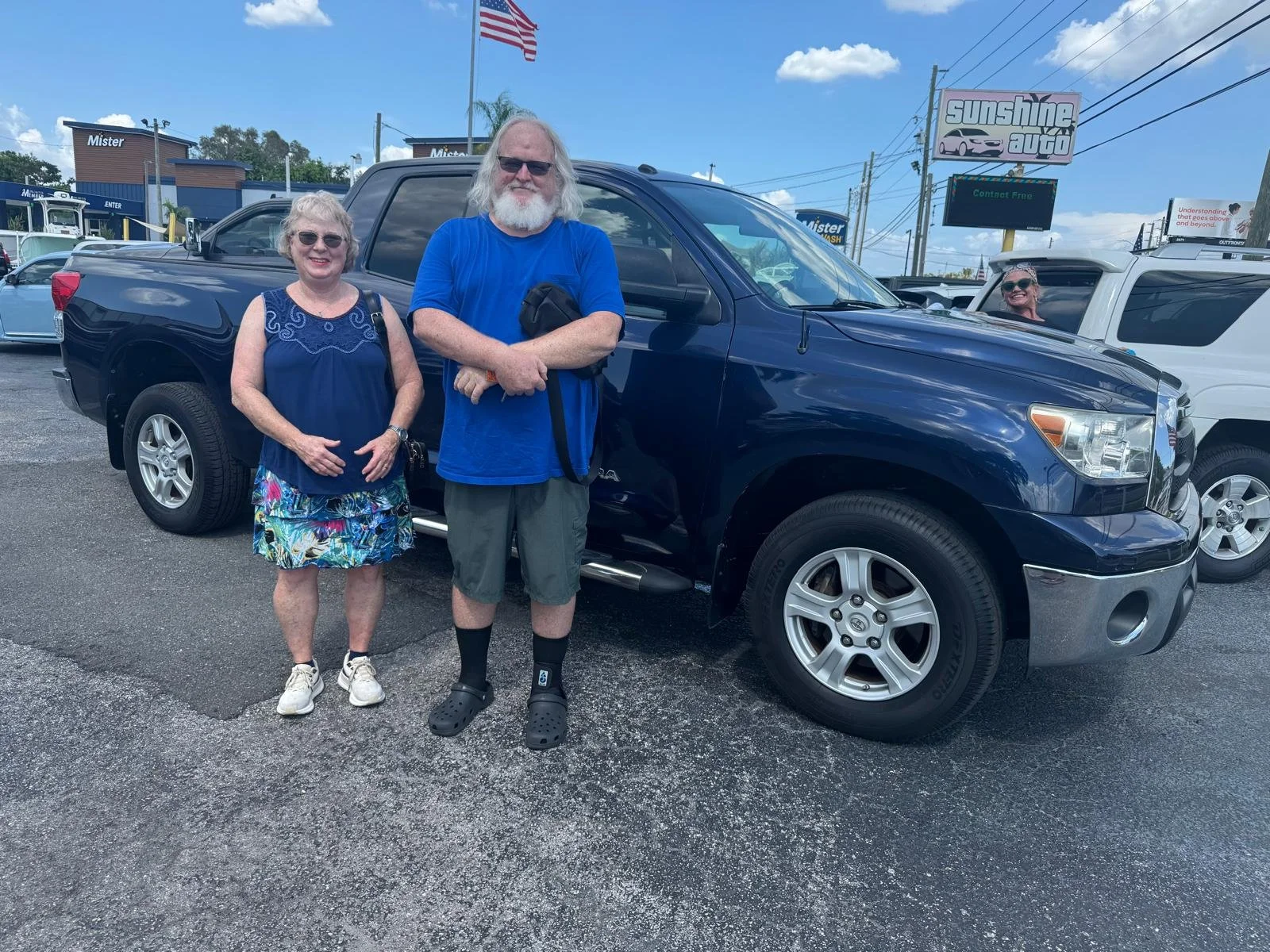 Two older women and a man standing in front of a black pickup truck at a car dealership, with one woman smiling, the man with sunglasses and a beard, and the other woman peeking from behind a white vehicle, all under a partly cloudy sky.