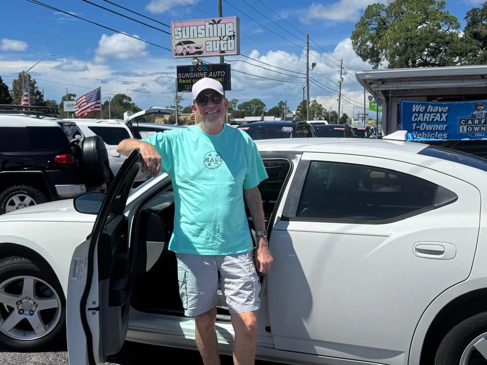 A man wearing sunglasses, a white cap, a aqua T-shirt, and white shorts standing next to a white car with the driver's door open at a car dealership lot on a sunny day.