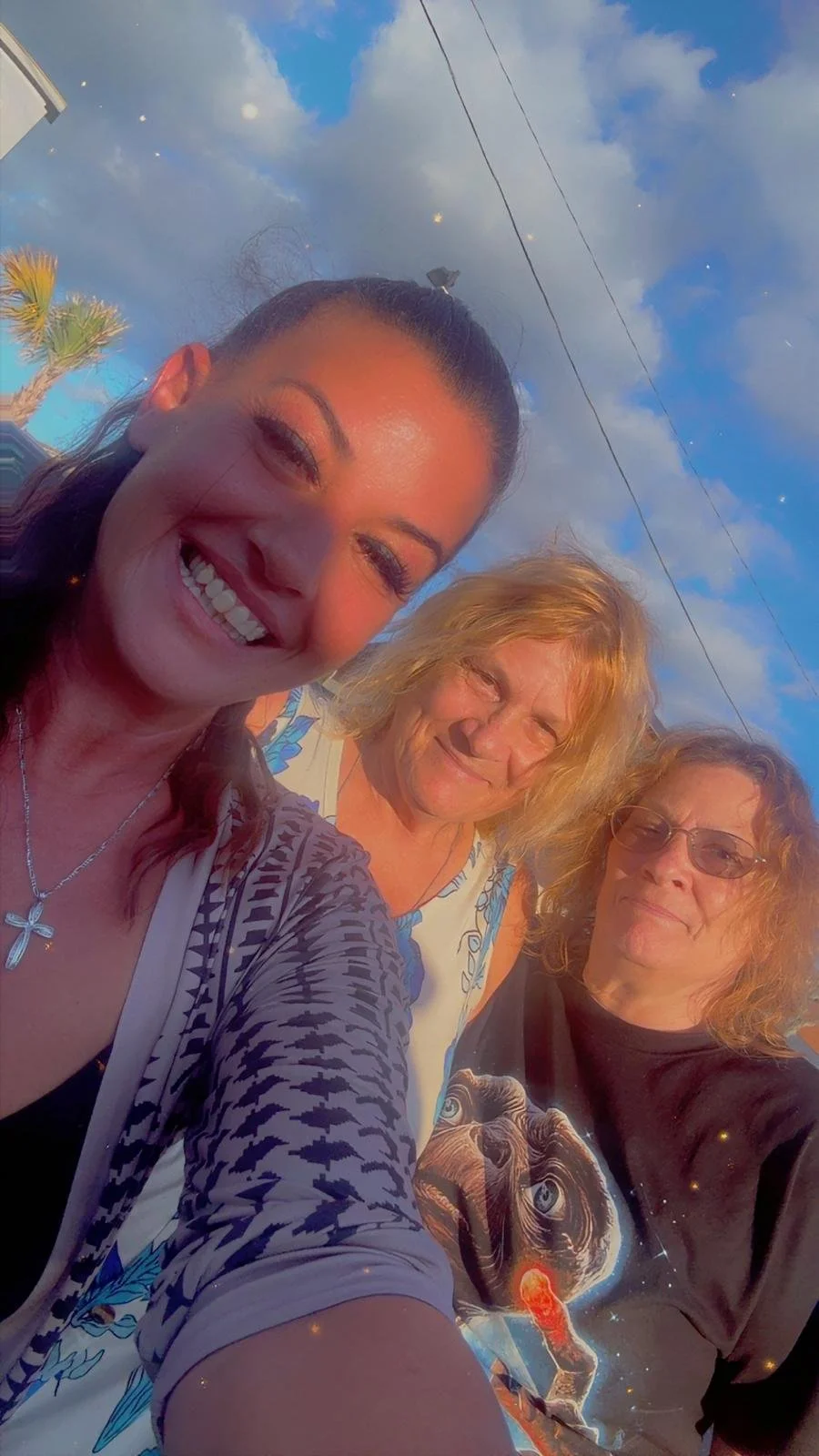 Three women taking a selfie outdoors in the late afternoon or early evening, with a partly cloudy sky and power lines in the background. The woman on the left has dark hair, is smiling broadly, and wears a necklace with a cross. The middle woman has 