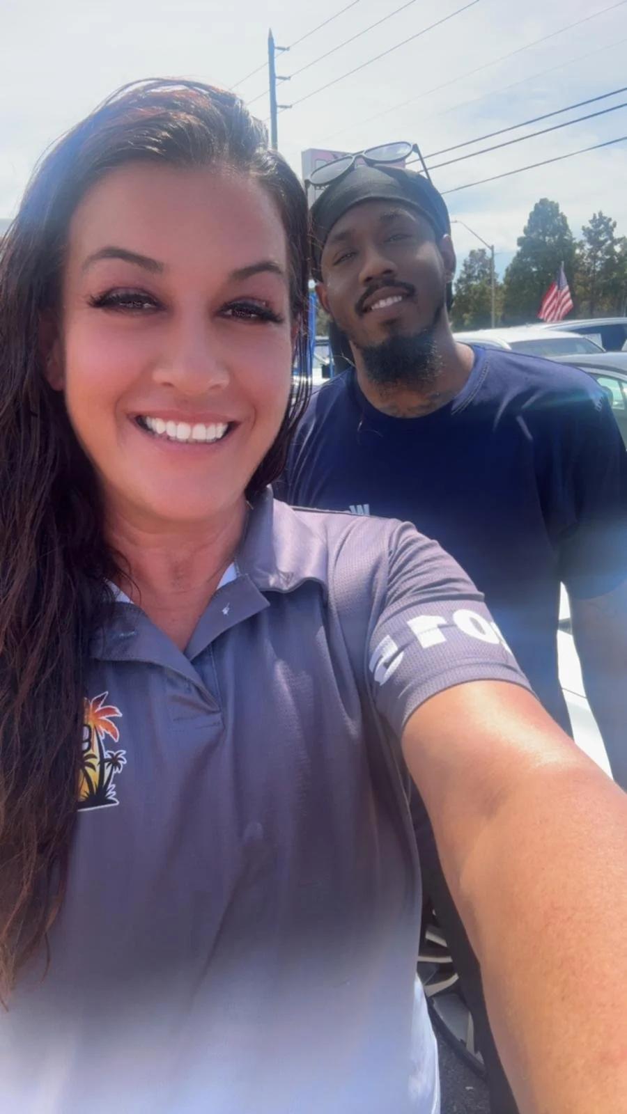 Two people taking a selfie outdoors on a sunny day, with cars and power lines in the background. The woman in the foreground has long dark hair and is wearing a gray shirt with a logo, smiling at the camera. The man behind her has a beard, glasses on