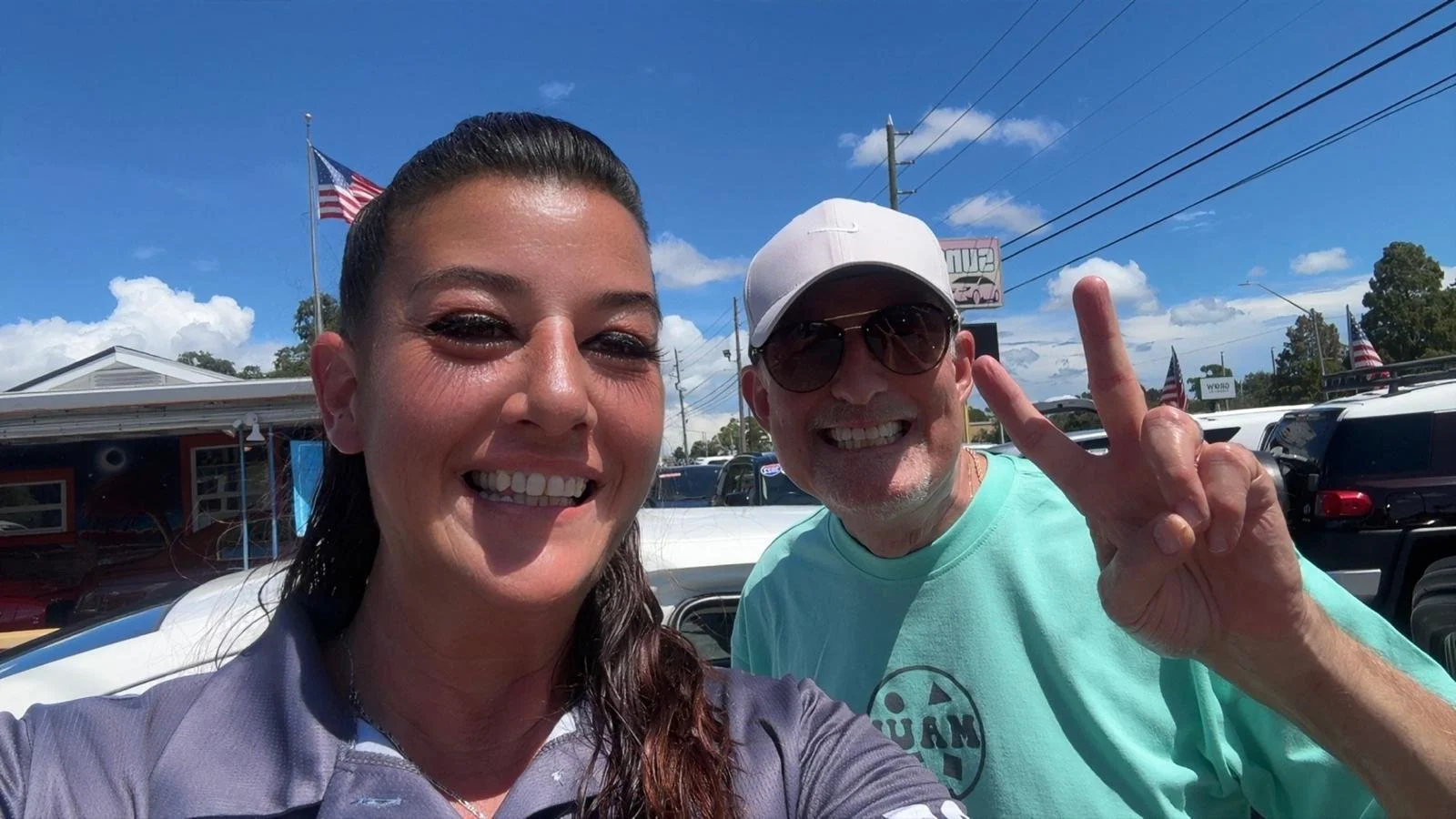 Smiling woman with dark hair and man wearing white cap and sunglasses, making a peace sign, in a parking lot on a sunny day with American flags and a building in the background.