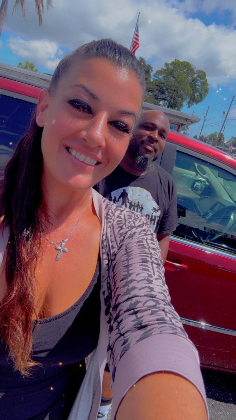 A woman taking a selfie with a man in front of a red vehicle outdoors, with trees, a house, and an American flag in the background under a partly cloudy sky.