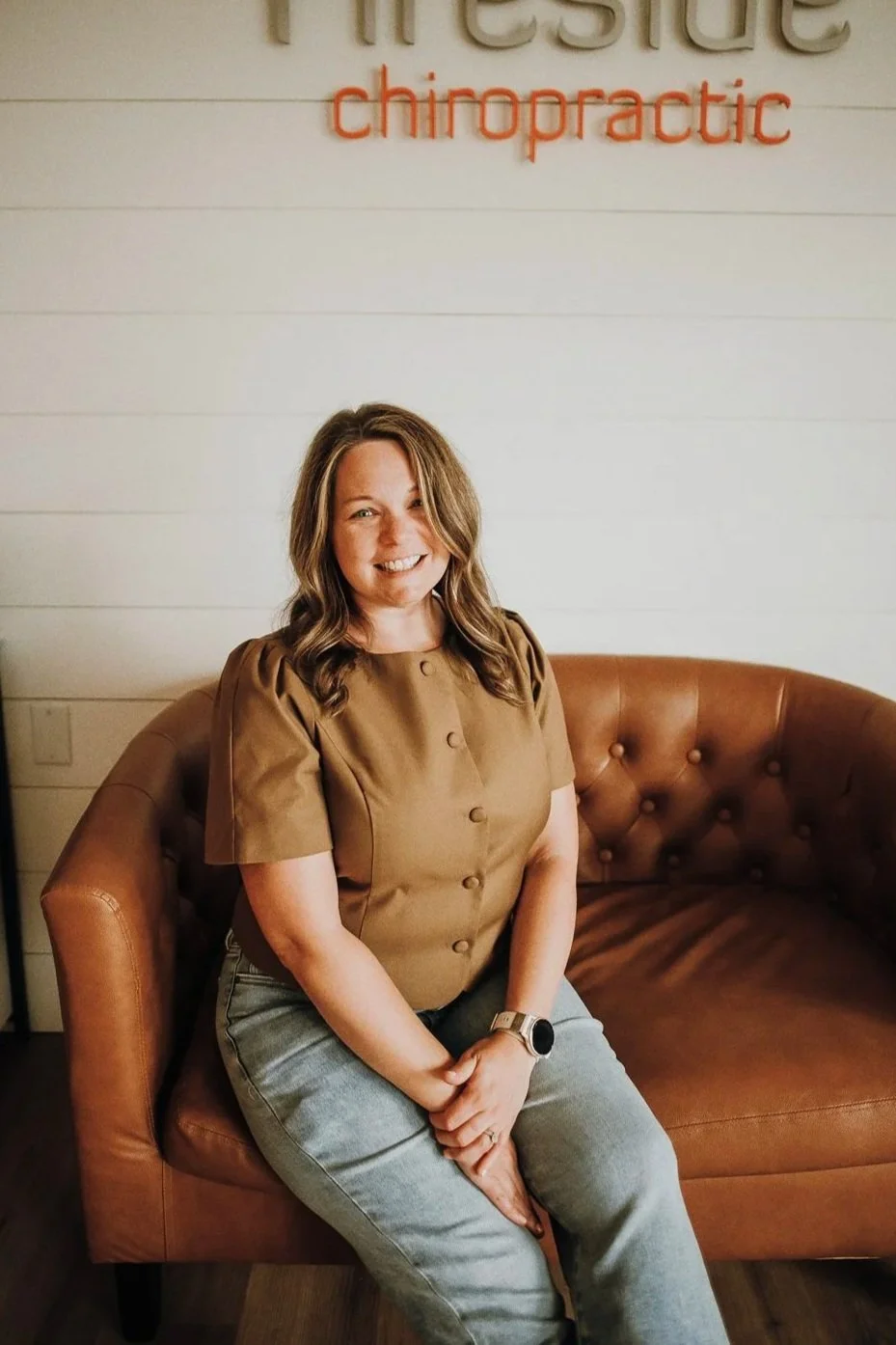 A smiling woman with long brown hair wearing a white Fireside Chiropractic sweatshirt, standing at a reception desk with a modern wooden front panel, in a chiropractic office.