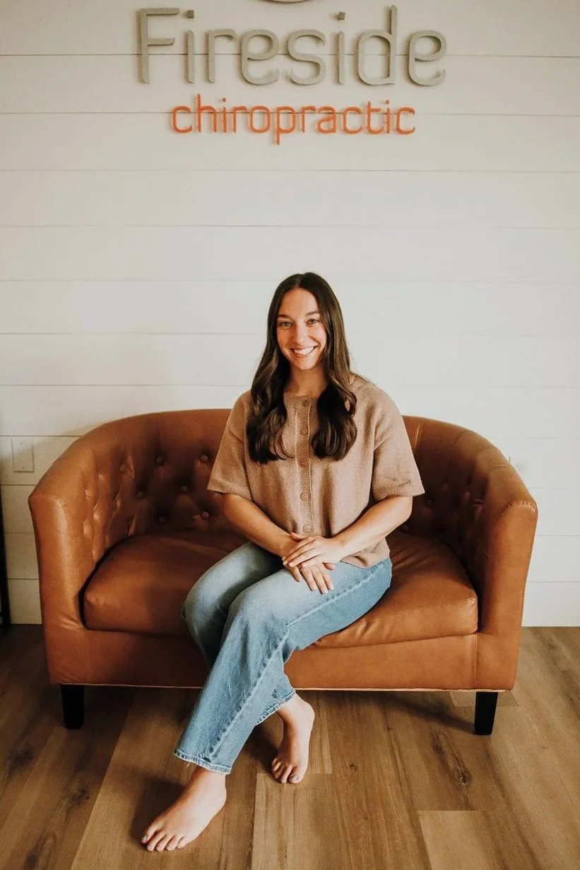 A woman with long dark hair smiling while sitting on a brown leather couch against a plain white wall, wearing a white high-neck shirt with a logo for "Fire" and "chiropractic".