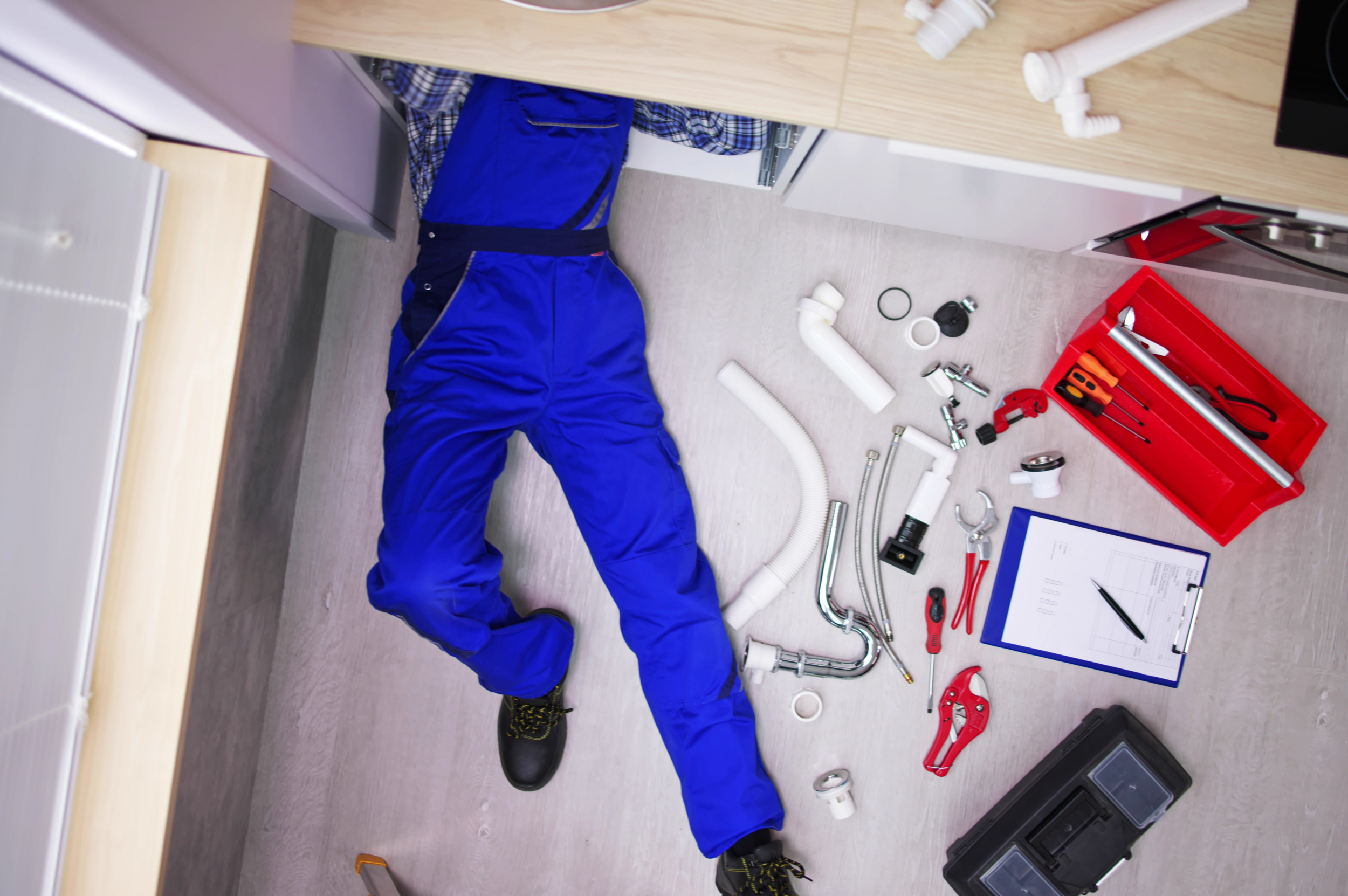 A plumber lying on the floor in a kitchen, surrounded by plumbing tools and pipes, including a red toolbox, pipe clamps, a pipe wrench, screwdrivers, a clipboard with papers, and various fittings.