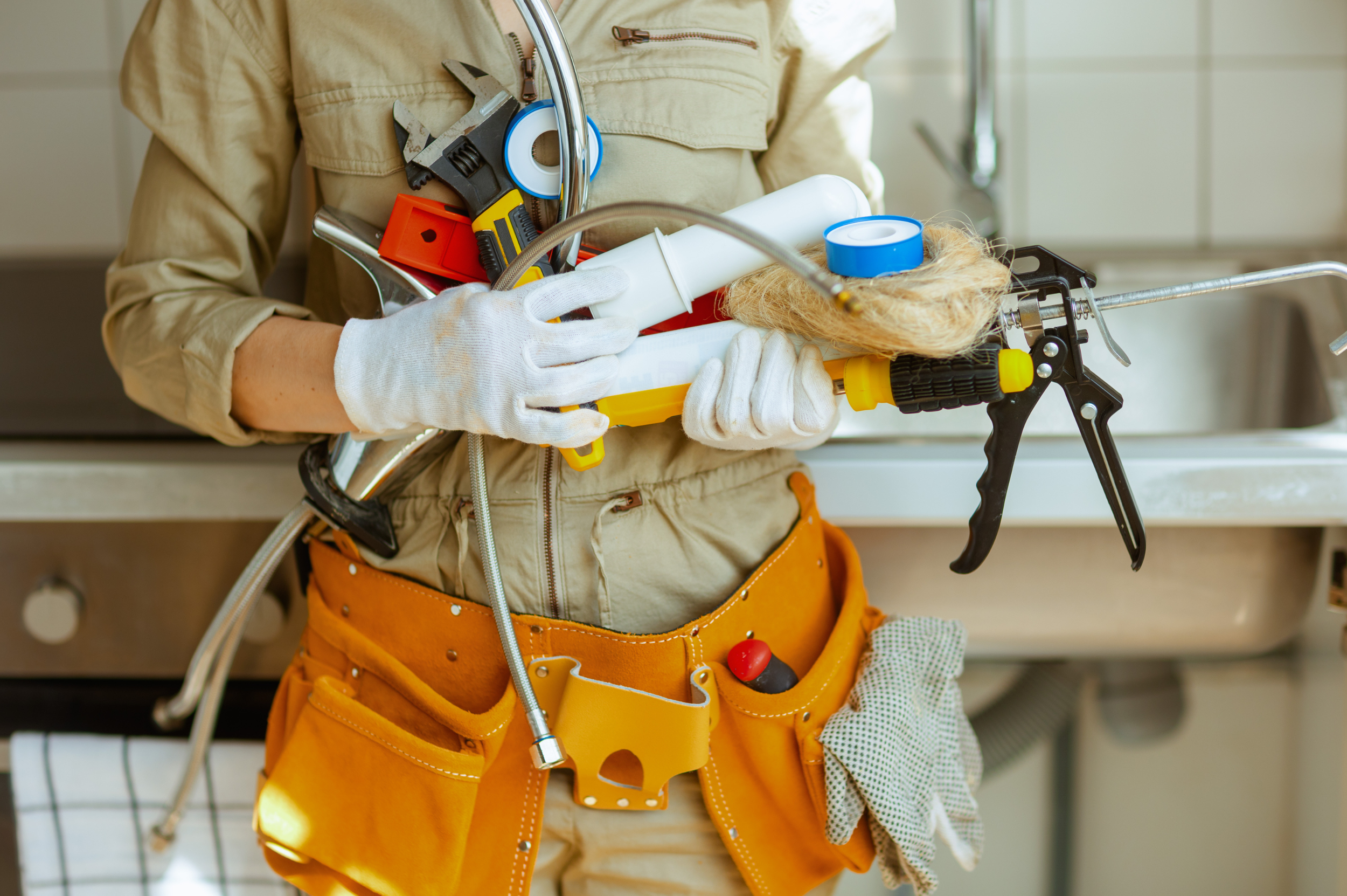 Plumber in a beige shirt and orange tool belt holding various plumbing tools and supplies, including a wrench, plunger, pipe, and tape.