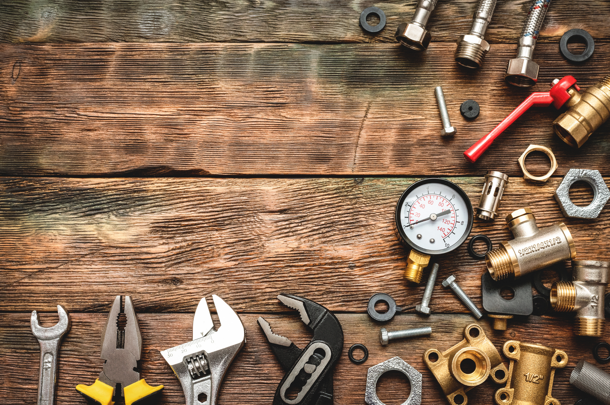 Various plumbing tools and fittings arranged on a wooden surface, including wrenches, adjustable pliers, a pressure gauge, nuts, bolts, and pipe connectors.