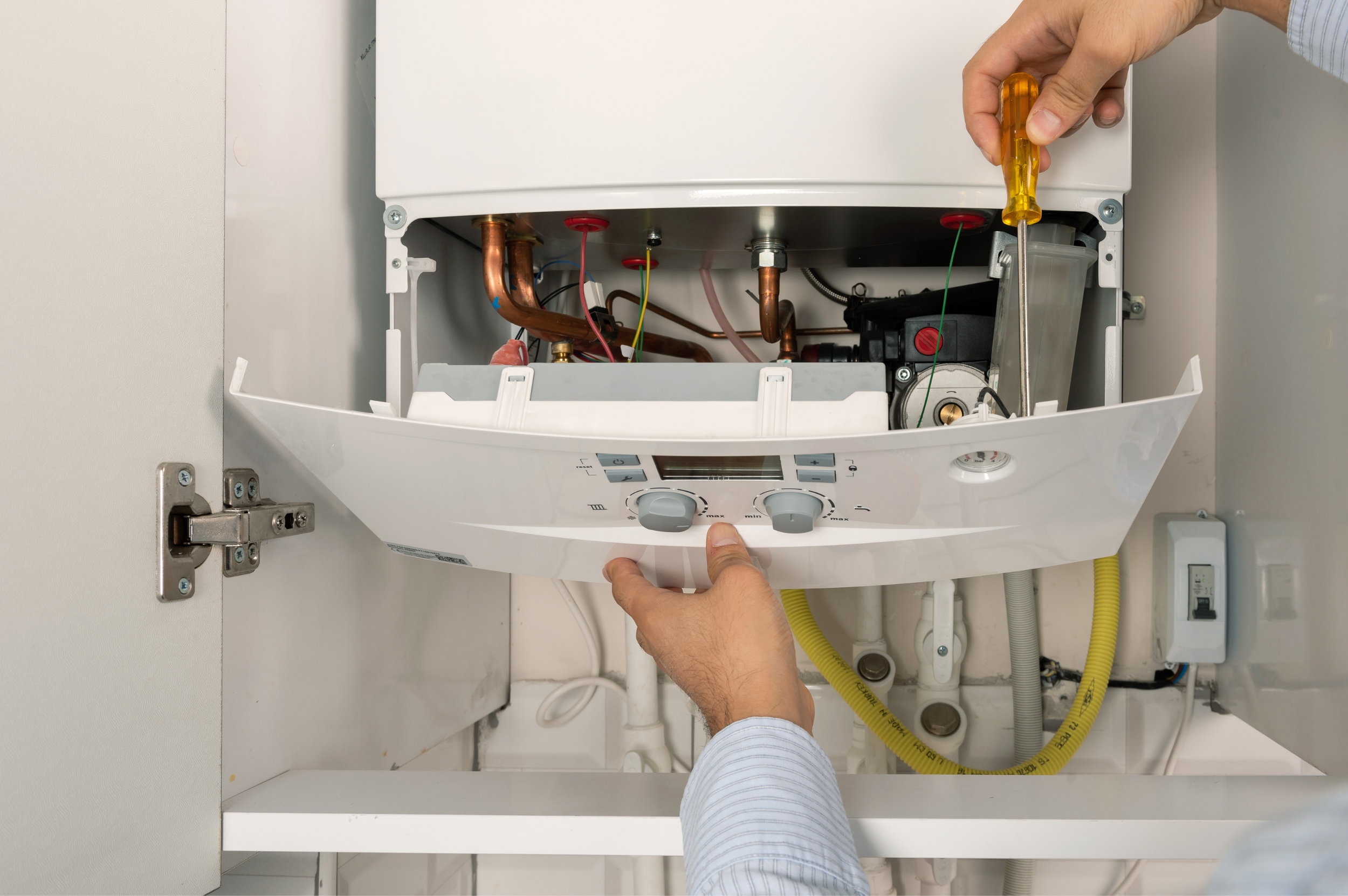 A plumber holding a screwdriver working inside a wall-mounted water heater, showing internal components and copper pipes.