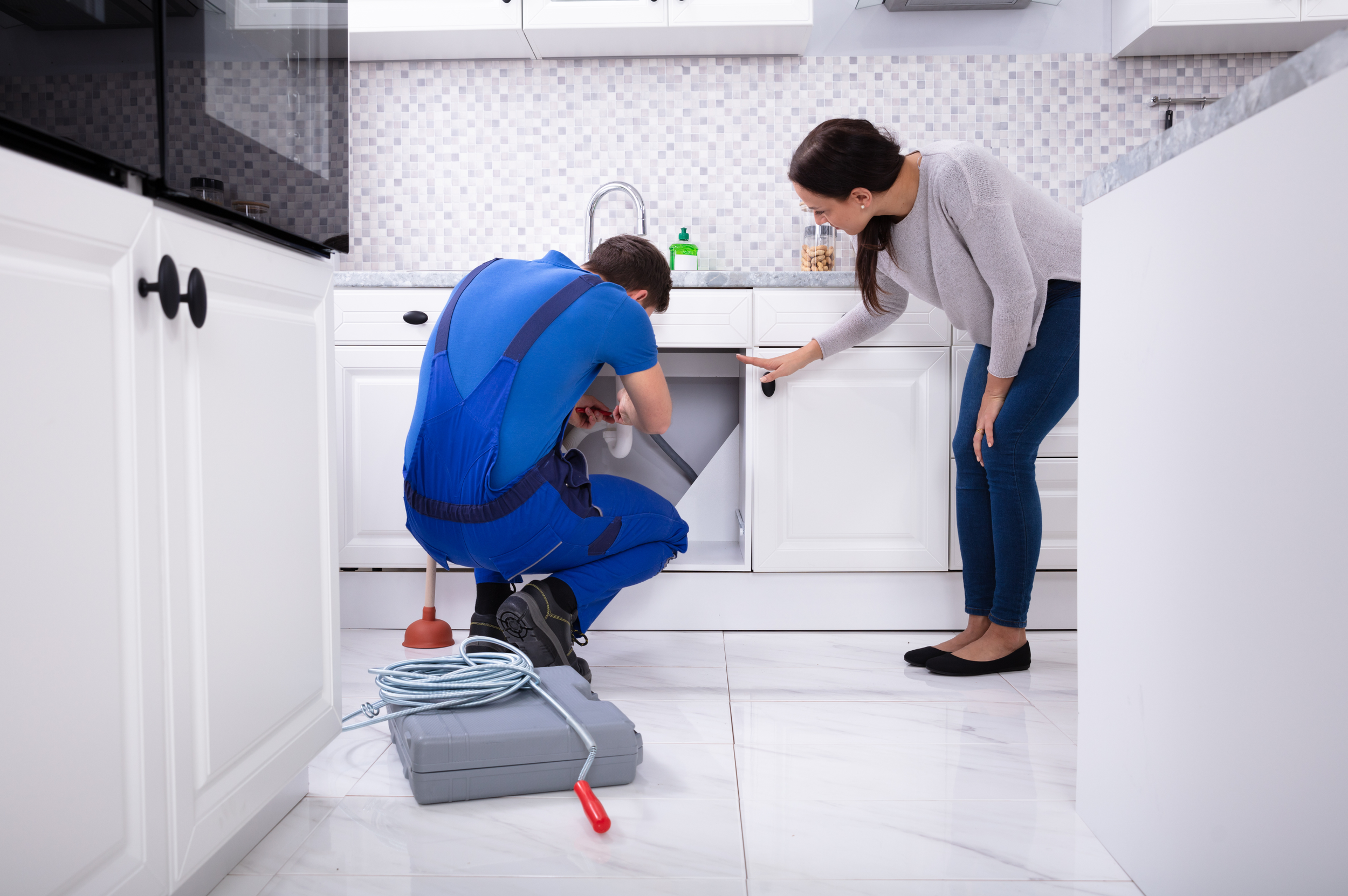 A plumber repairing a kitchen sink while a customer looks on, with plumbing tools on the floor.