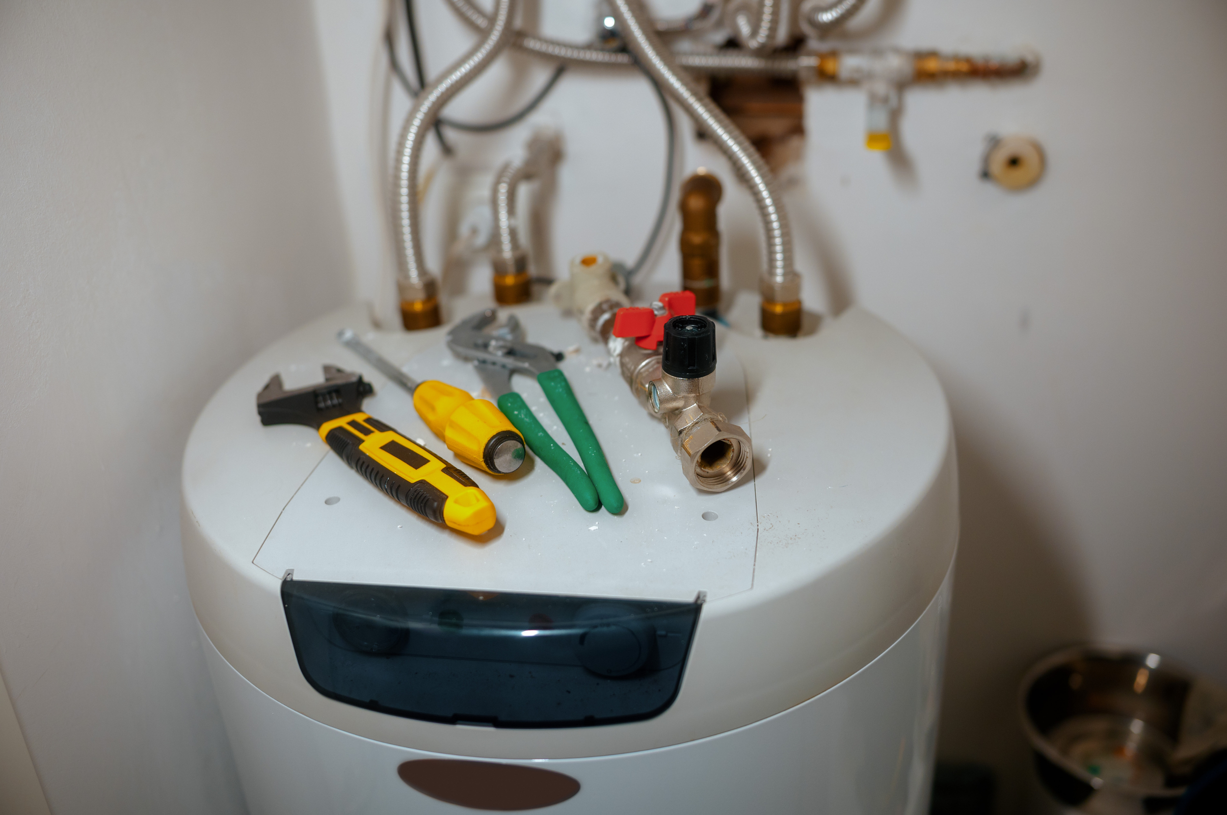 Tools and plumbing parts on top of a water heater, including screwdrivers, pliers, and pipes, with a partially open panel in the background.