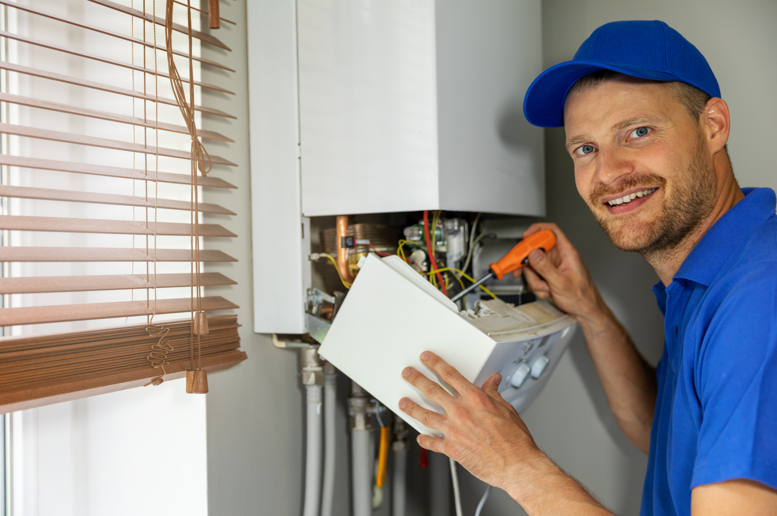 A plumber in a blue cap and shirt fixing a boiler near a window with blinds.