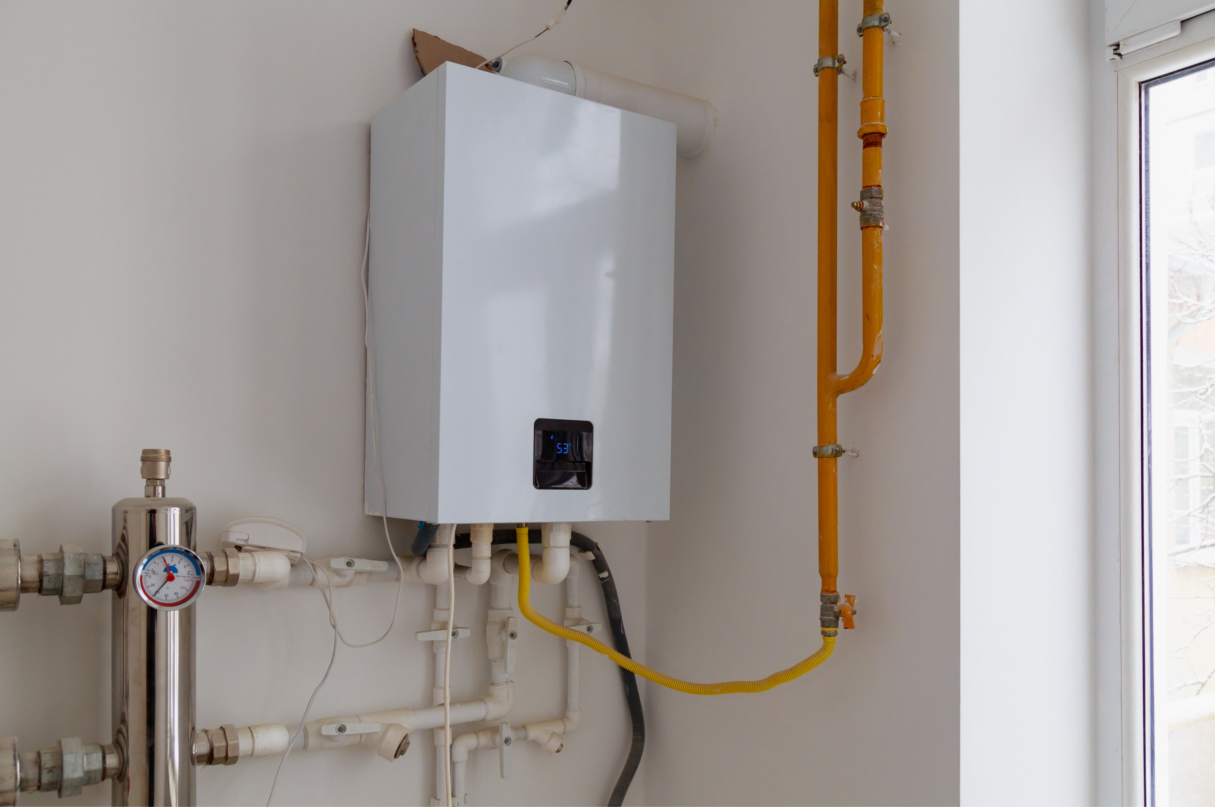 A white wall-mounted boiler with a digital display. Pipes of various sizes and colors, including orange, yellow, and white, are connected to the boiler and running vertically and horizontally along the wall. A window with a snowy outdoor scene is partially visible on the right.