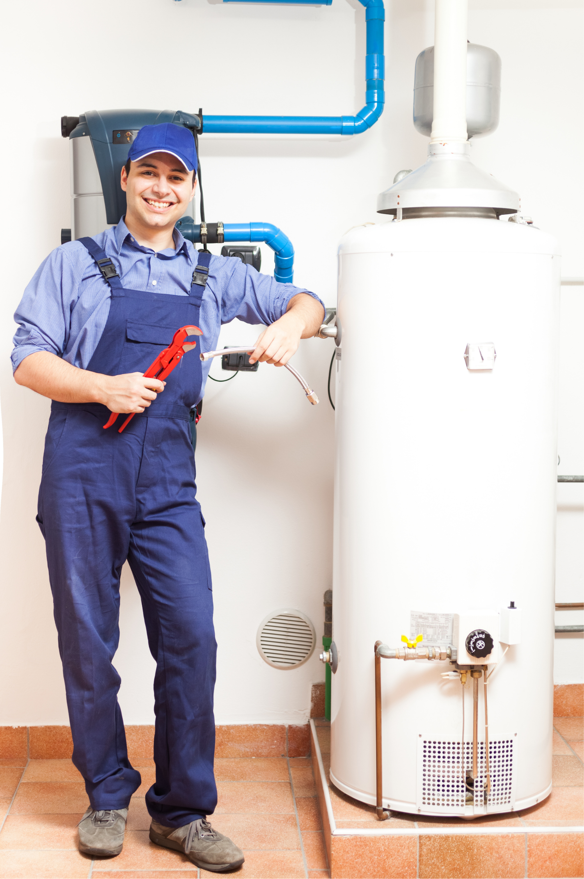 Plumber wearing a blue cap, blue scrubs, and gray shoes standing in a room with tiled floors, smiling while working on a white water heater, holding a red pipe wrench in his right hand.