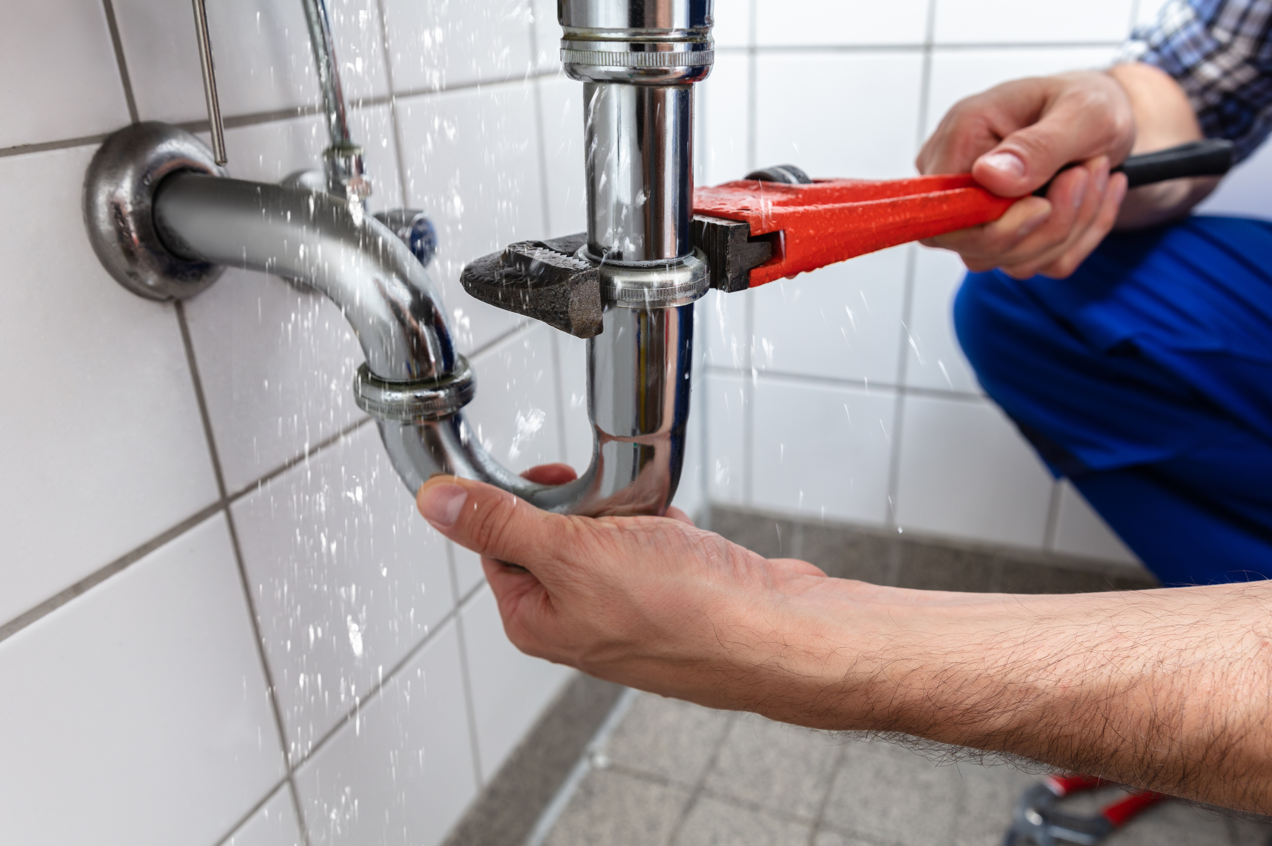 A plumber using a pipe wrench to fix or install a copper pipe under a sink with water dripping.