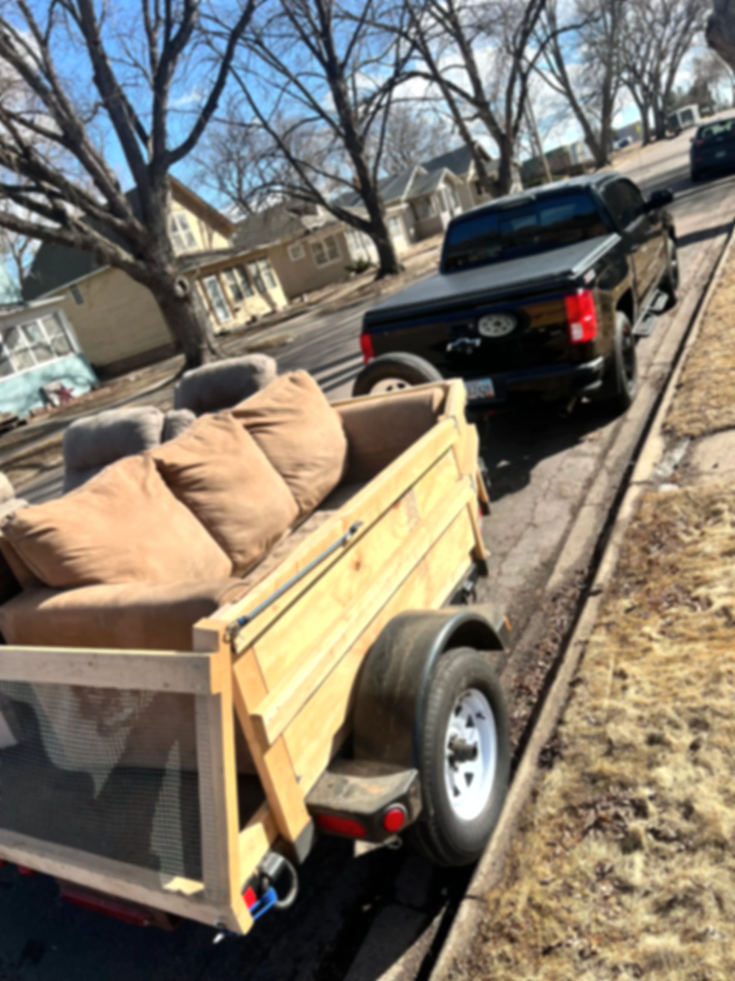 A black pickup truck parked on the street with a trailer attached, carrying furniture and a pet crate, in a neighborhood with leafless trees and houses.
