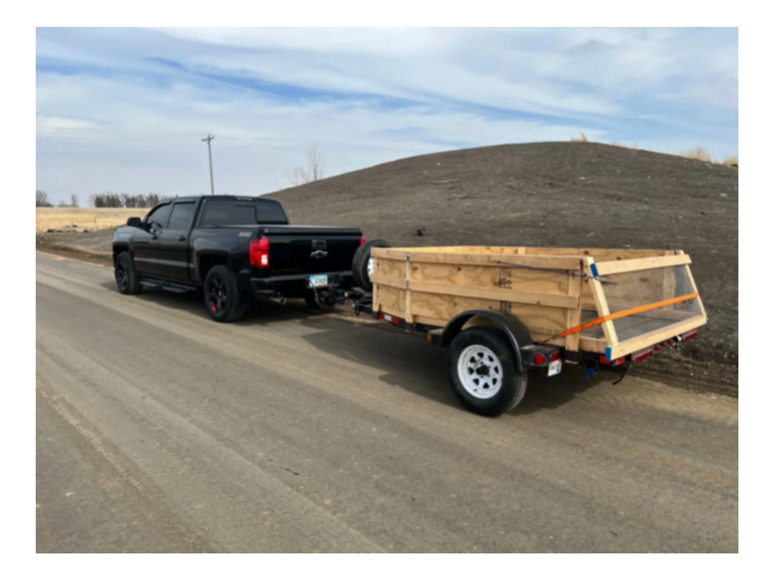 A black Chevrolet Silverado pickup truck pulling a small wooden trailer on a country road next to a large bare dirt hill.