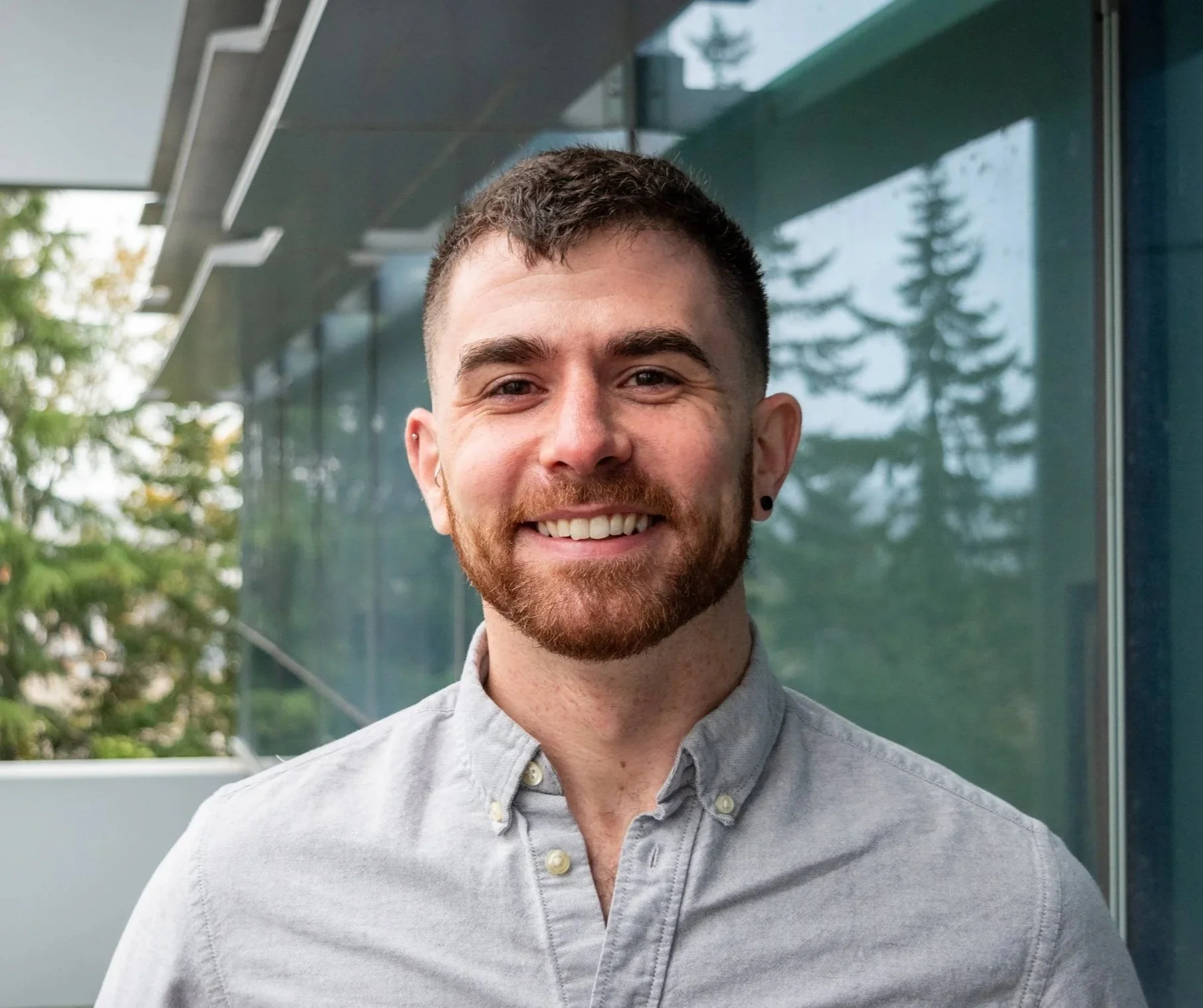 Close-up of a young man with a beard smiling, wearing a light gray button-up shirt, standing outdoors with trees and a glass building in the background.