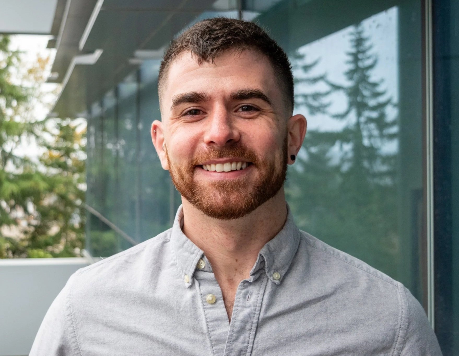 A young man with short brown hair and a beard is smiling at the camera. He is wearing a light gray button-up shirt. The background shows a building with large glass windows and some trees outside.