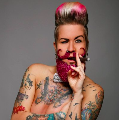 A drag king (male impersonator) with pink and blonde hair styled in an updo, holding a cigar with a glittery pink glove, and covering his mouth with his hand, showing tattoos on his arms and chest.