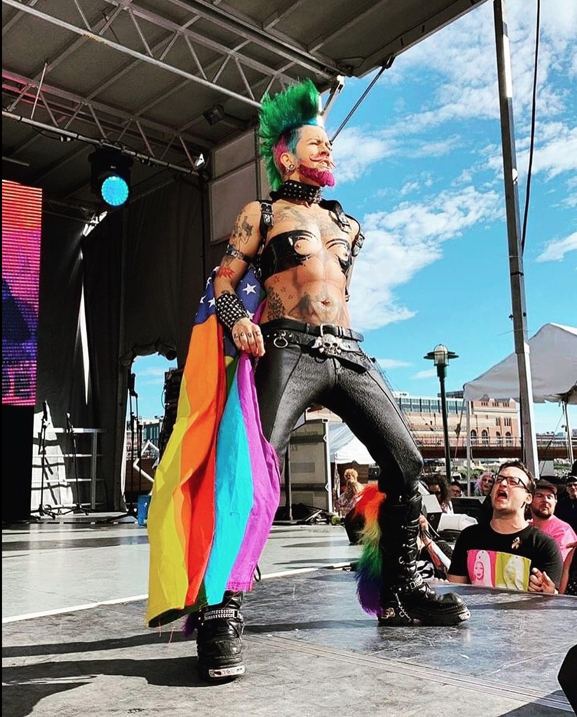 A person with a punk style on stage, holding a rainbow pride flag, wearing black pants, boots, and studded accessories, with colorful hair and tattoos, performing in front of an outdoor audience on a sunny day.
