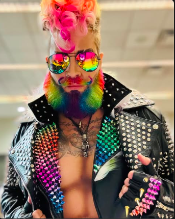 Person with colorful dyed beard, rainbow-colored curly hair, rainbow sunglasses, and a studded leather jacket.