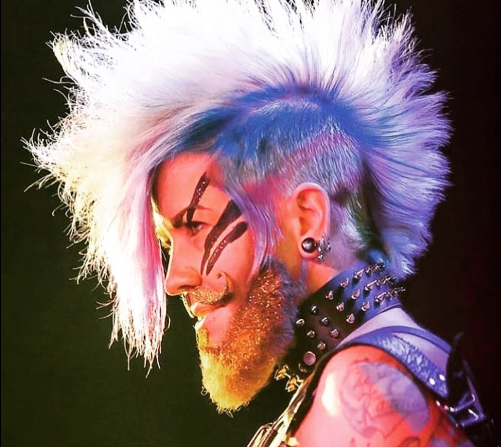 Close-up of a person with spiked, multicolored hair, face and neck tattoos, wearing black earrings, and a spiked choker, smiling sideways.