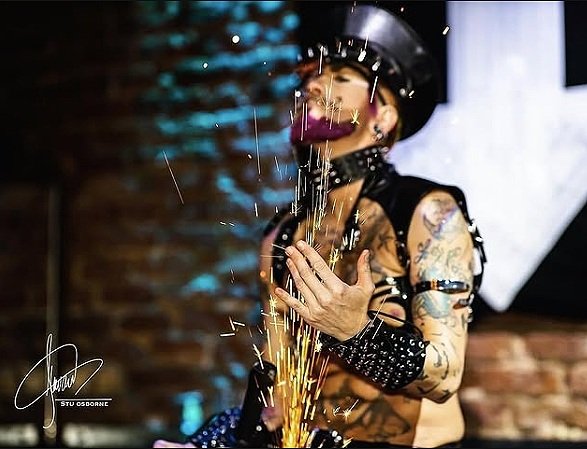 A drag king dressed in a leather and studded outfit wearing a hat and sunglasses, with tattoos on arms, is sitting with eyes closed and a smile on face while sparks are flying around them