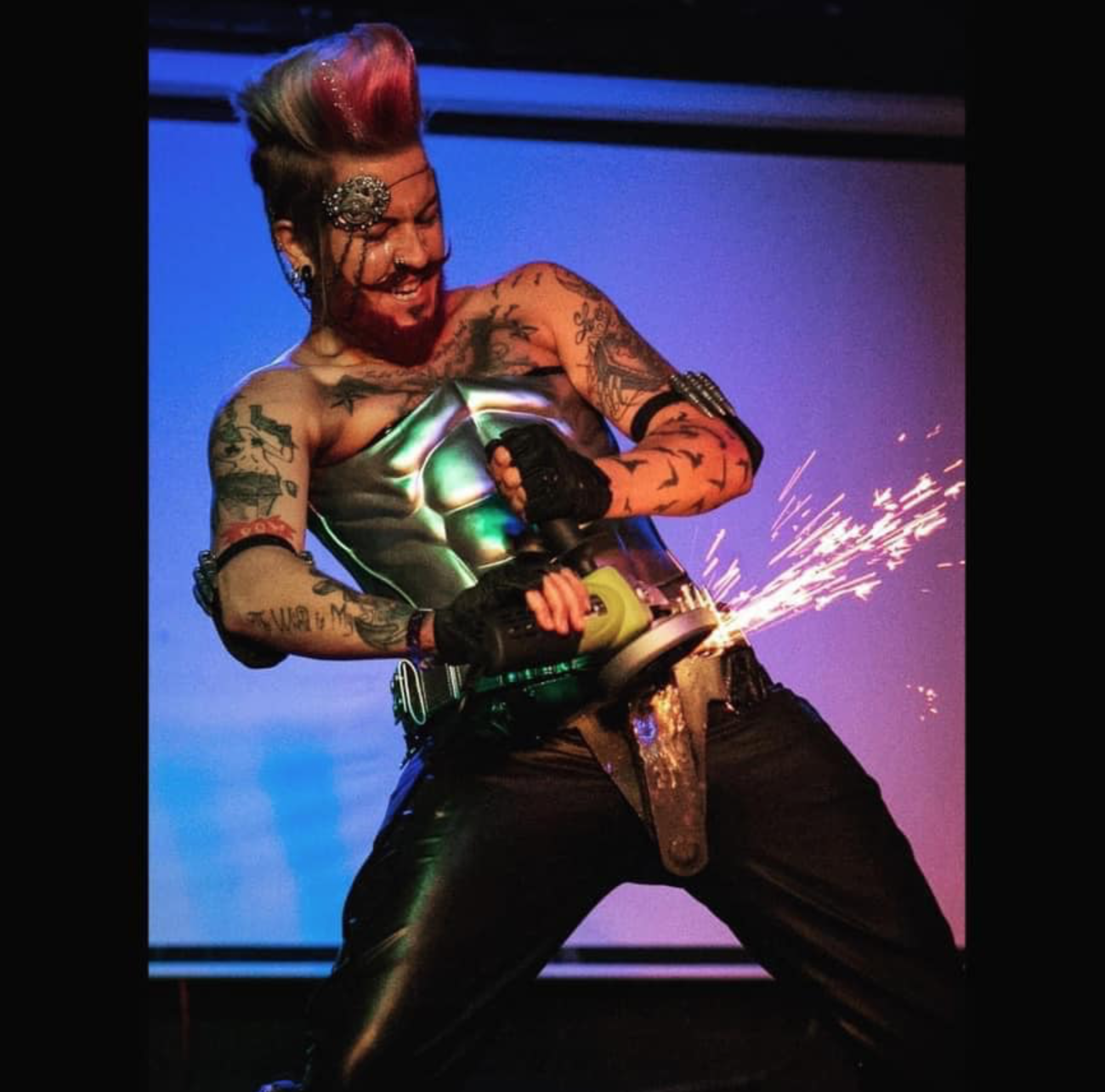 Person with tattoos and colorful hair using an angle grinder, sparks flying, smiling, in a performance setting.