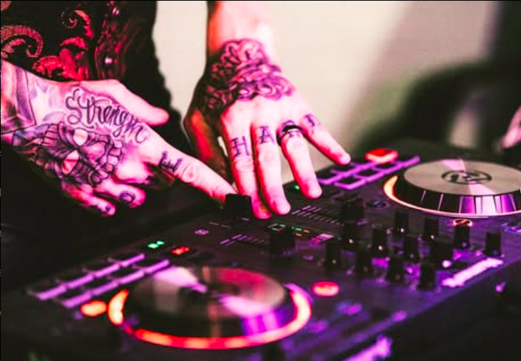 Close-up of tattooed hands using a DJ mixer, with colorful lights on the equipment.