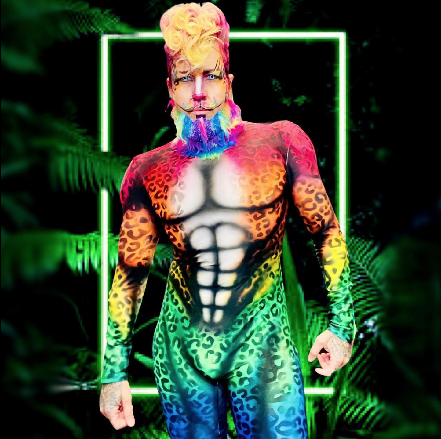 Person with rainbow-colored beard and stylized rainbow hair, wearing a body suit painted with a muscular human torso and animal print patterns, in front of a neon-green square frame and tropical foliage.