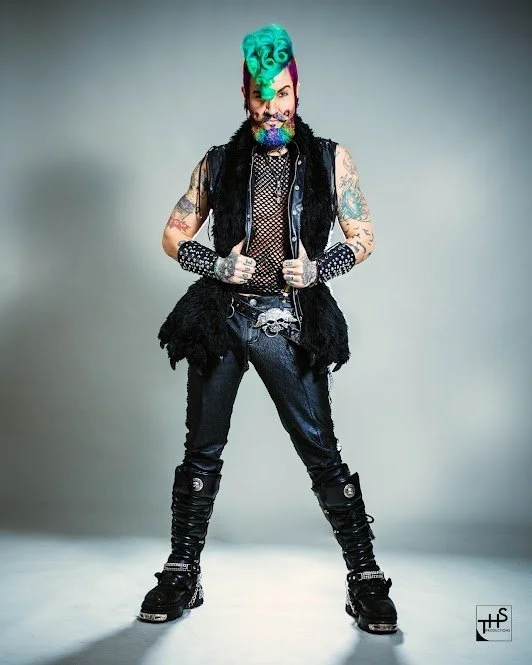 A man with bright turquoise hair styled in a vintage waves, an elaborate beard, and multiple tattoos. He is wearing a black furry vest, fishnet shirt, studded gloves, black pants with knee straps, and black boots, standing confidently with legs apart against a plain background.