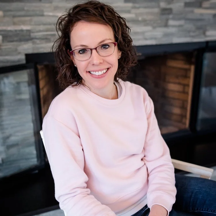 A woman with shoulder-length blonde hair and blue eyes is smiling outdoors in a grassy area with trees and a body of water in the background. She is wearing a dark green top with floral-patterned sleeves, a beaded necklace, and earrings.