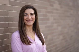 A woman with curly brown hair, glasses, and a light pink sweatshirt smiling while sitting indoors in front of a stone fireplace.