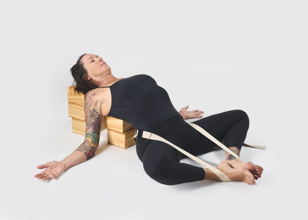 Woman in black workout clothes lying on the floor with a foam block under her back, using straps around her legs, in a studio with white background.