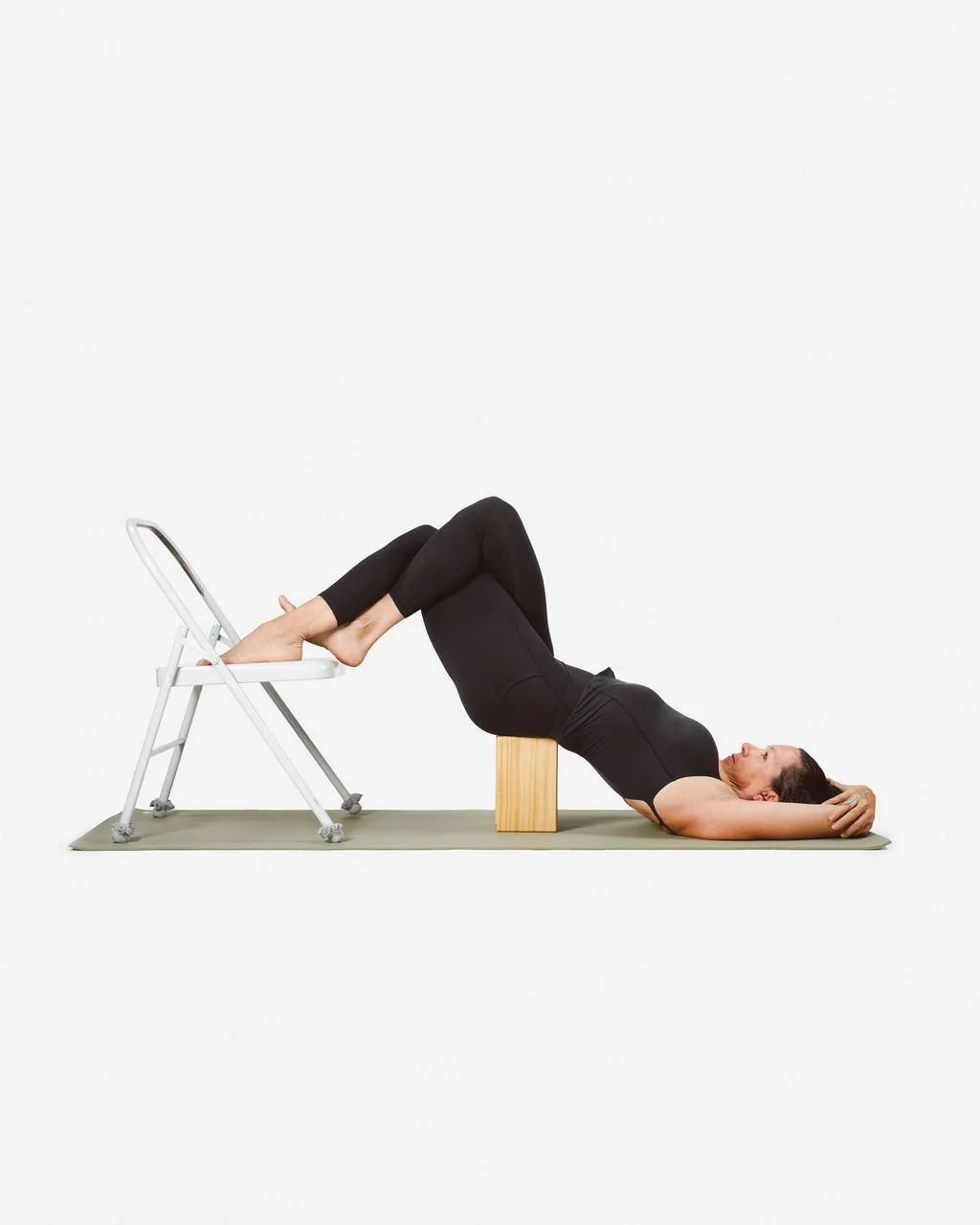 A woman practicing yoga in black activewear is doing a supported bridge pose with her feet on a chair and her hips on a wooden block, lying on a mat.