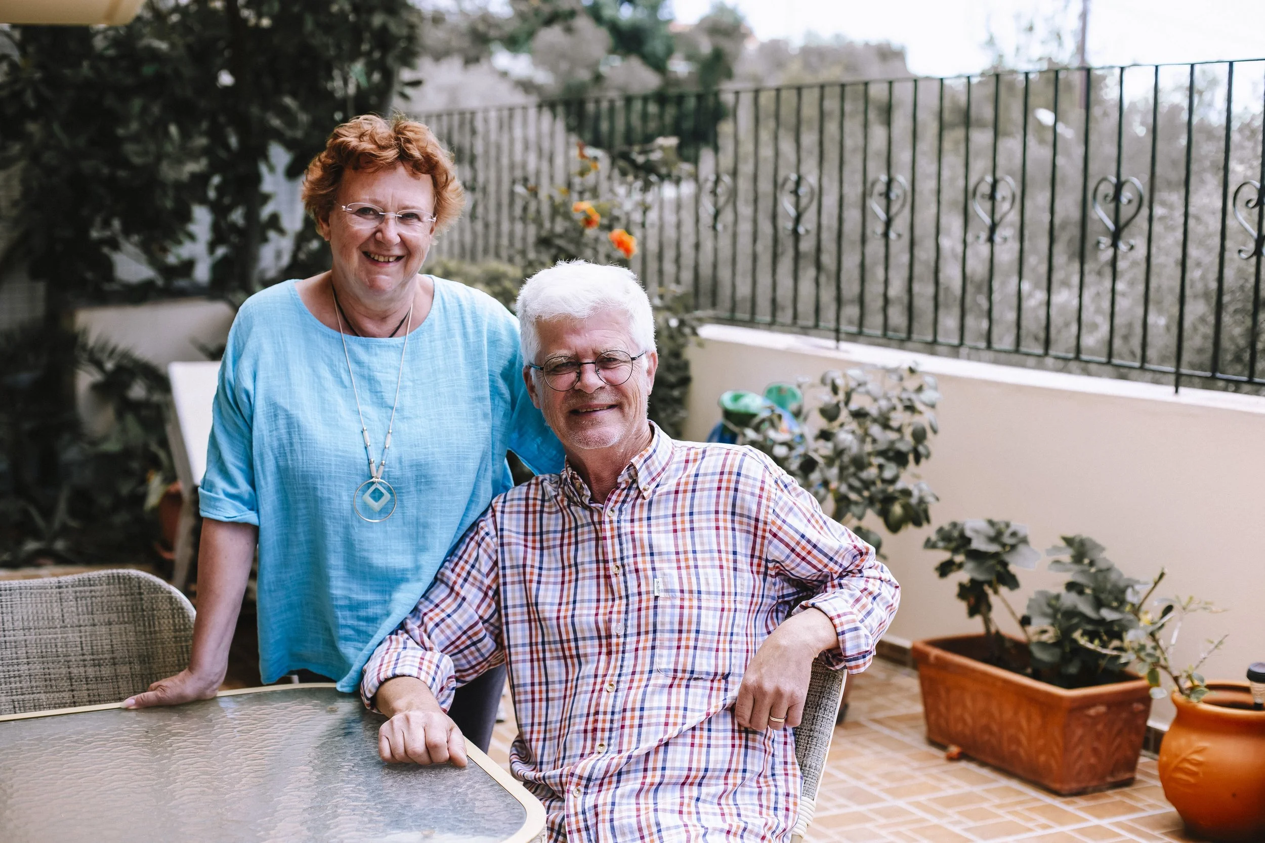 Kaksi ihmistä, nainen ja mies, istuvat ja seisovat terassilla, nauravat ja katsovat kameraan.
