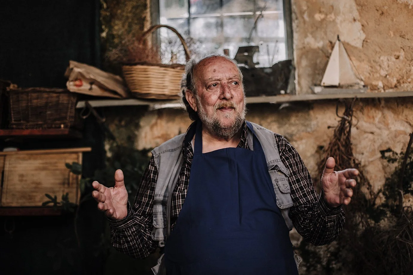 Vanha mies puhumassa Fabrica Farmin tiloissa, taustalla rouhea kiviseinä, jolla hylly ja keittiötarvikkeita