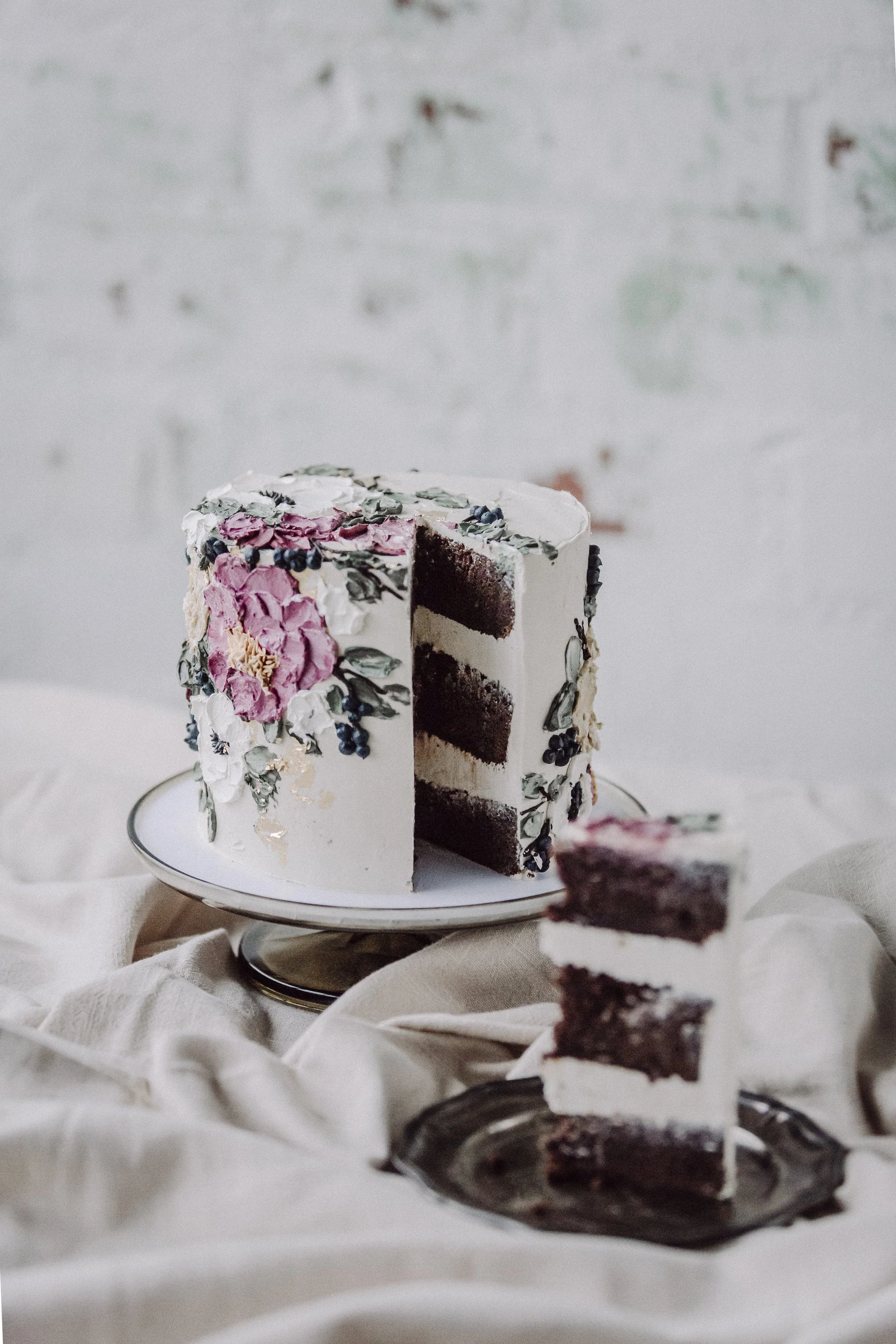 Koristeellinen kerroskakku, jonka päällä on vaaleanpunaisia ja vihreitä kukkia, ja jossa on kolme kerrosta suklaakakkua ja kerma-välitäytteitä, aseteltu valkoiselle tarjoilu­tiinalle, taustalla valkoinen seinä.