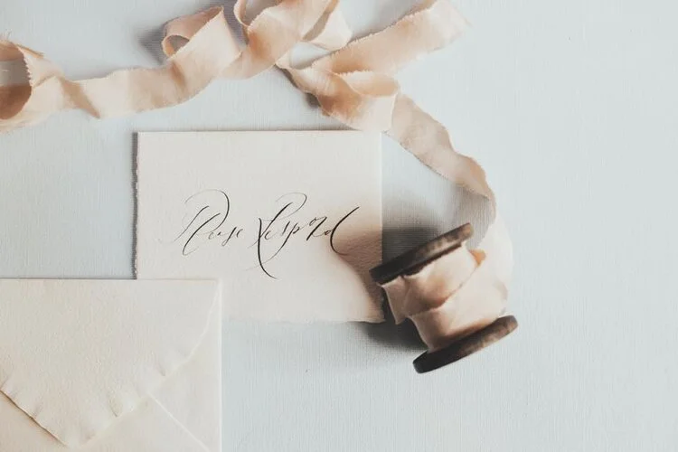 A piece of paper with cursive writing, a spool of ribbon, and an envelope on a light surface.