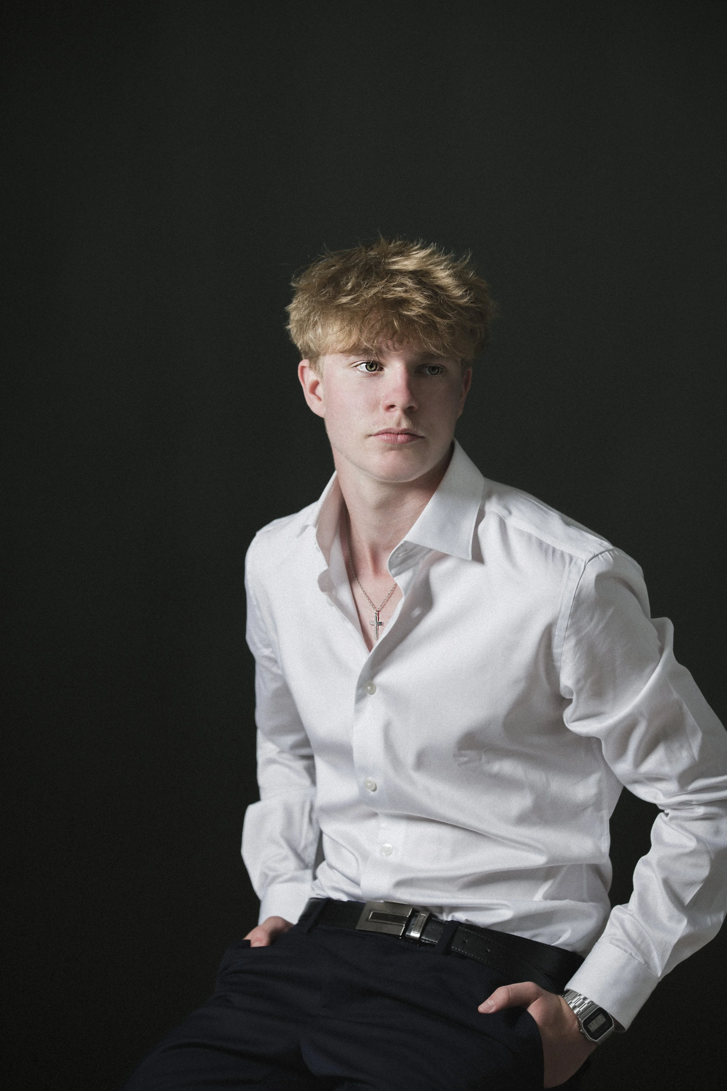 Nuori mies valkoinen paita ja rannekello, istuu pimeässä taustassa.