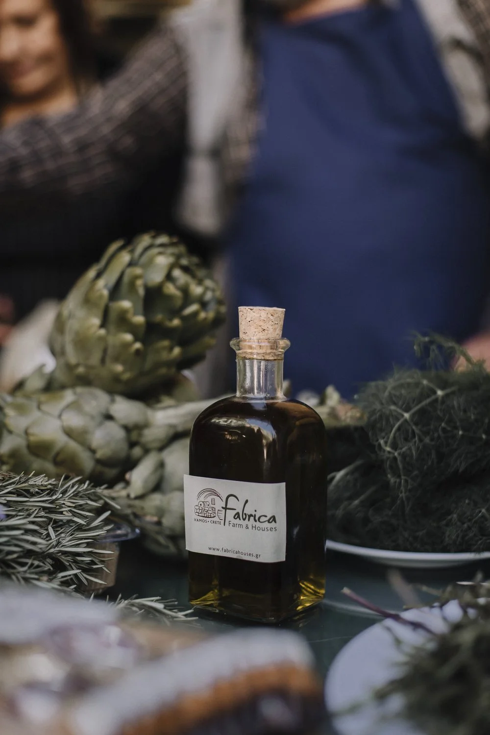 A bottle of olive oil labeled "Fabrica Farm & Houses" surrounded by artichokes, rosemary, and dill on a table.