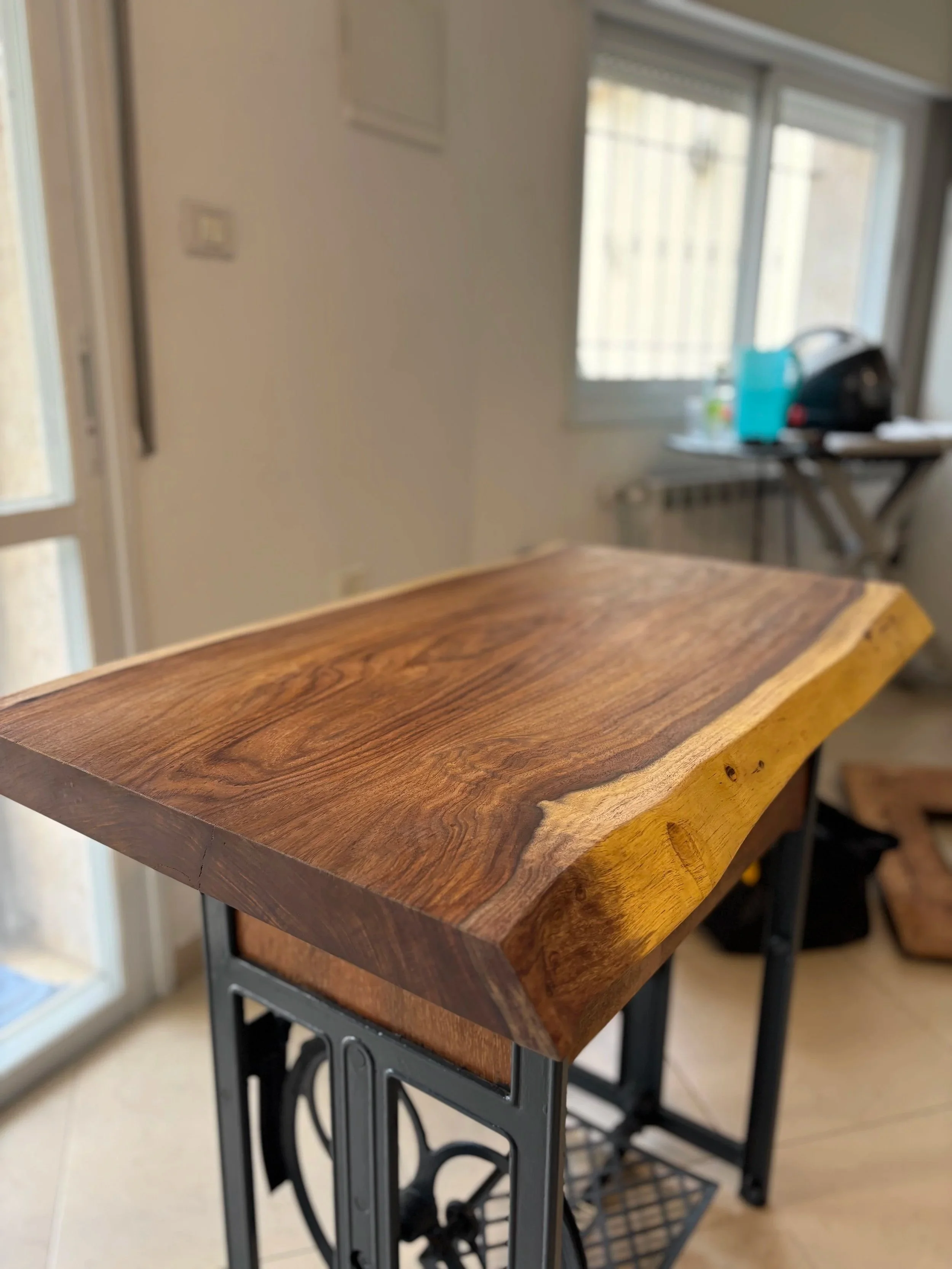 Close-up of a polished wooden table with a natural edge, mounted on a black metal stand, in a bright room with windows.