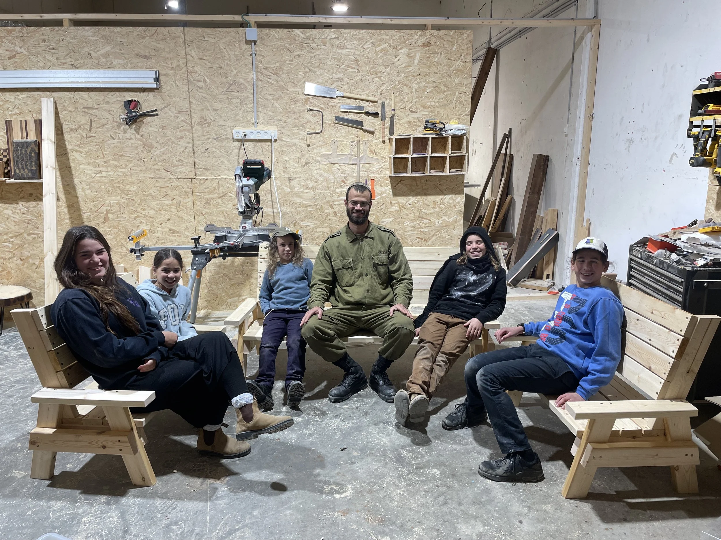 Group of six people, including children and an adult man in work clothes, sitting on wooden benches in a woodworking workshop.