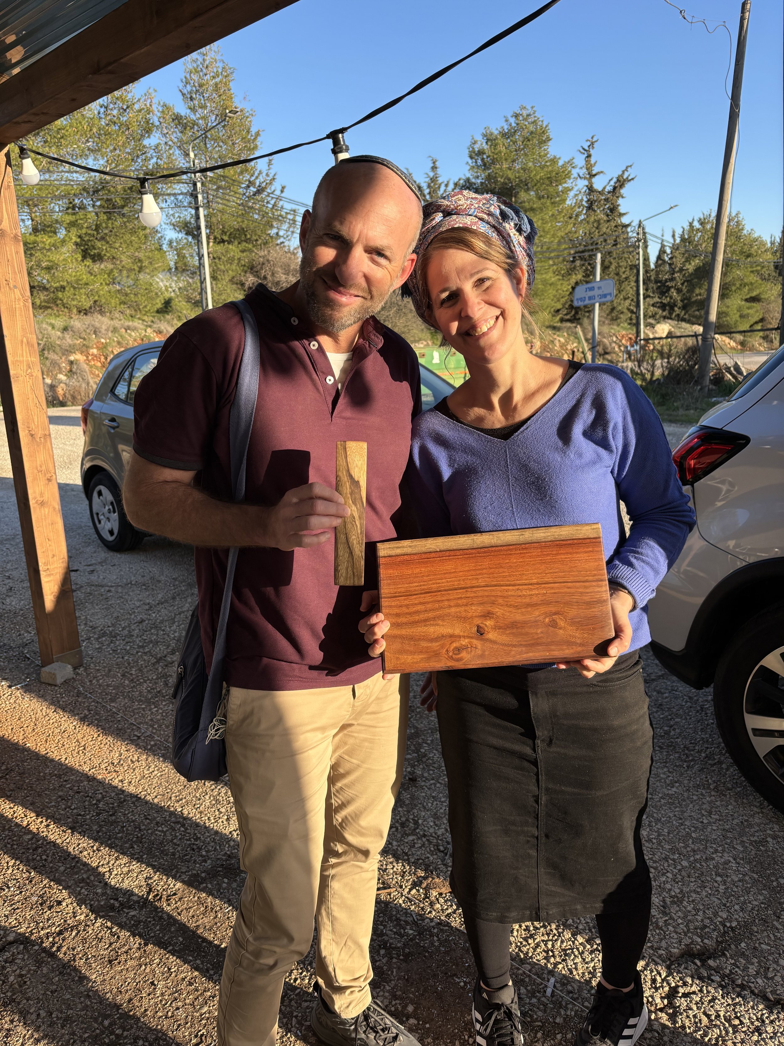 A man and a woman smiling outdoors, holding a wooden box, with cars and trees in the background, during sunny weather.
