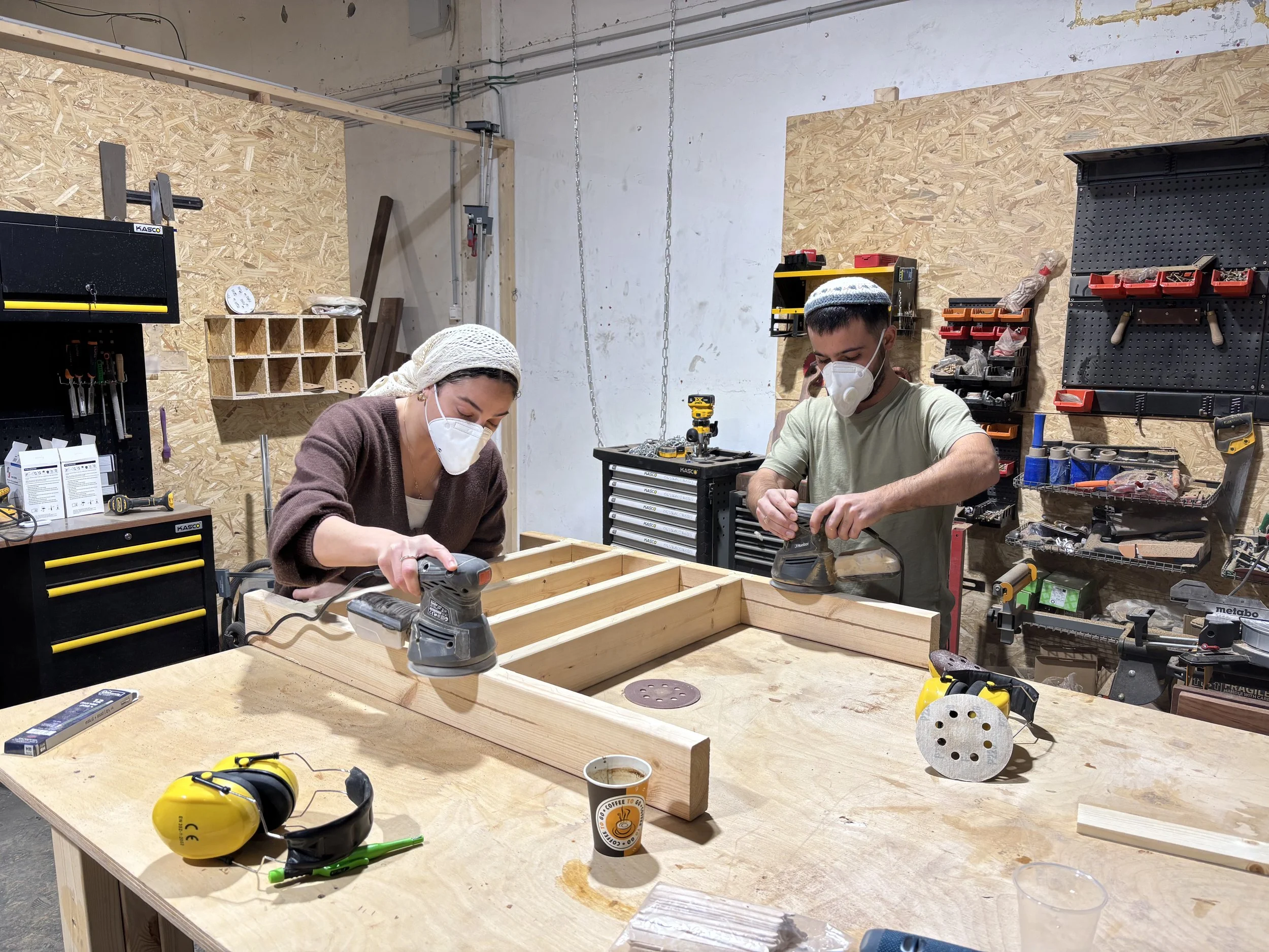 Two people wearing face masks working on a woodworking project in a workshop, using handheld power sanders on a wooden frame. The workshop has various tools and equipment on the walls and workbenches.