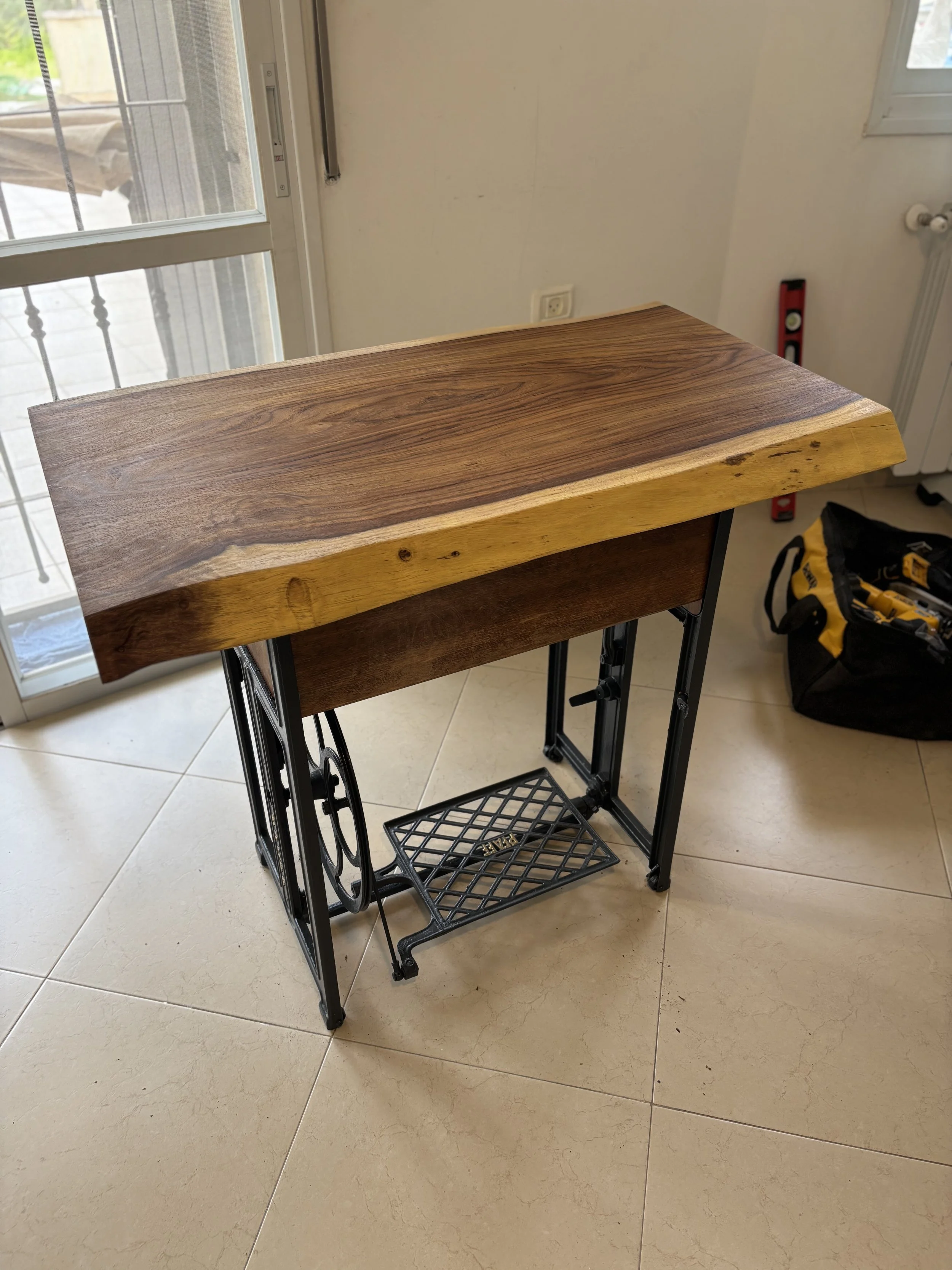 A vintage Singer sewing table with a wooden top in a room with beige tiled floor, a sliding glass door, and a bag of tools in the background.