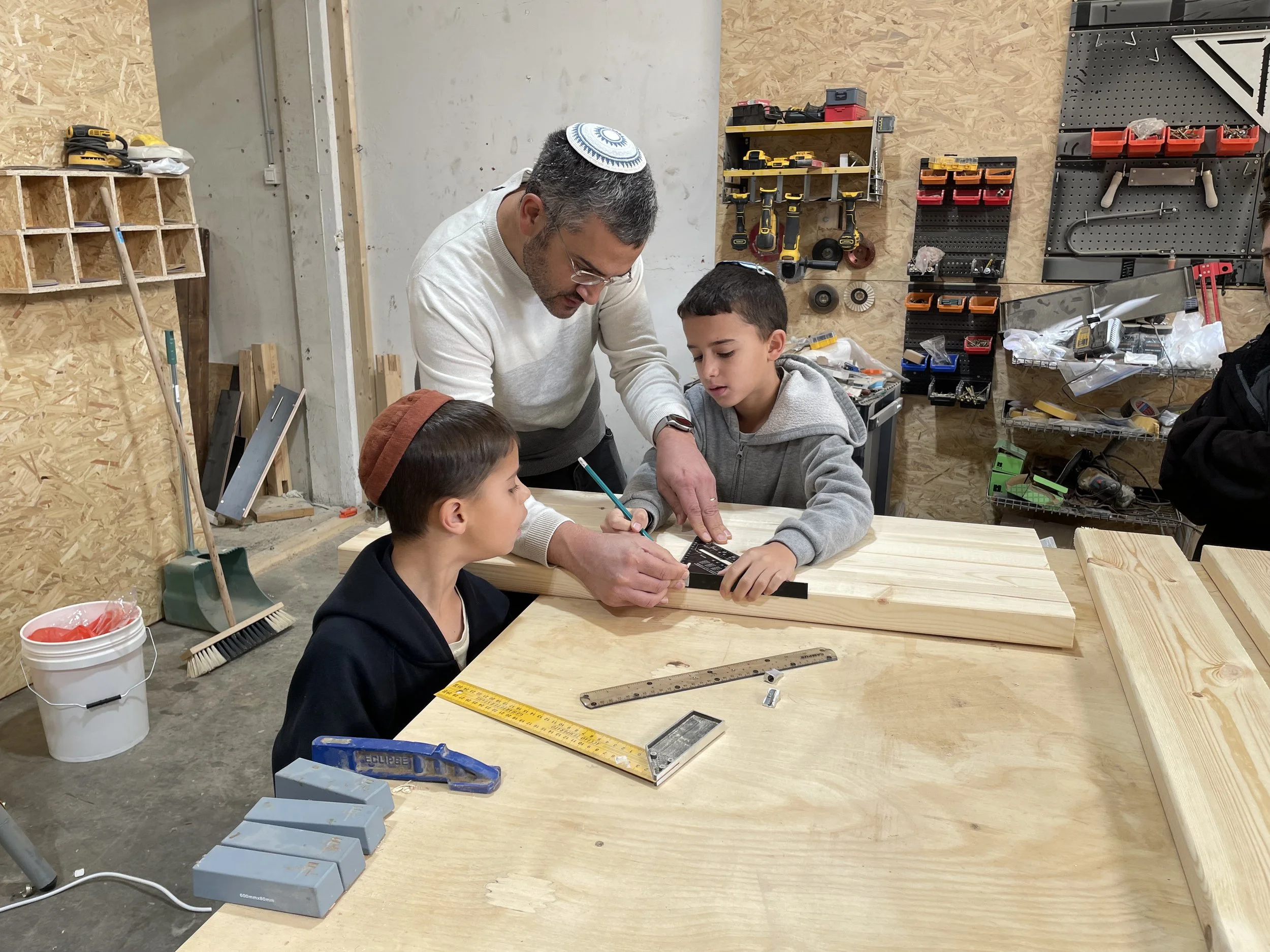 A man and two children working together on a woodworking project in a workshop, with hand tools and wood on the workbench.