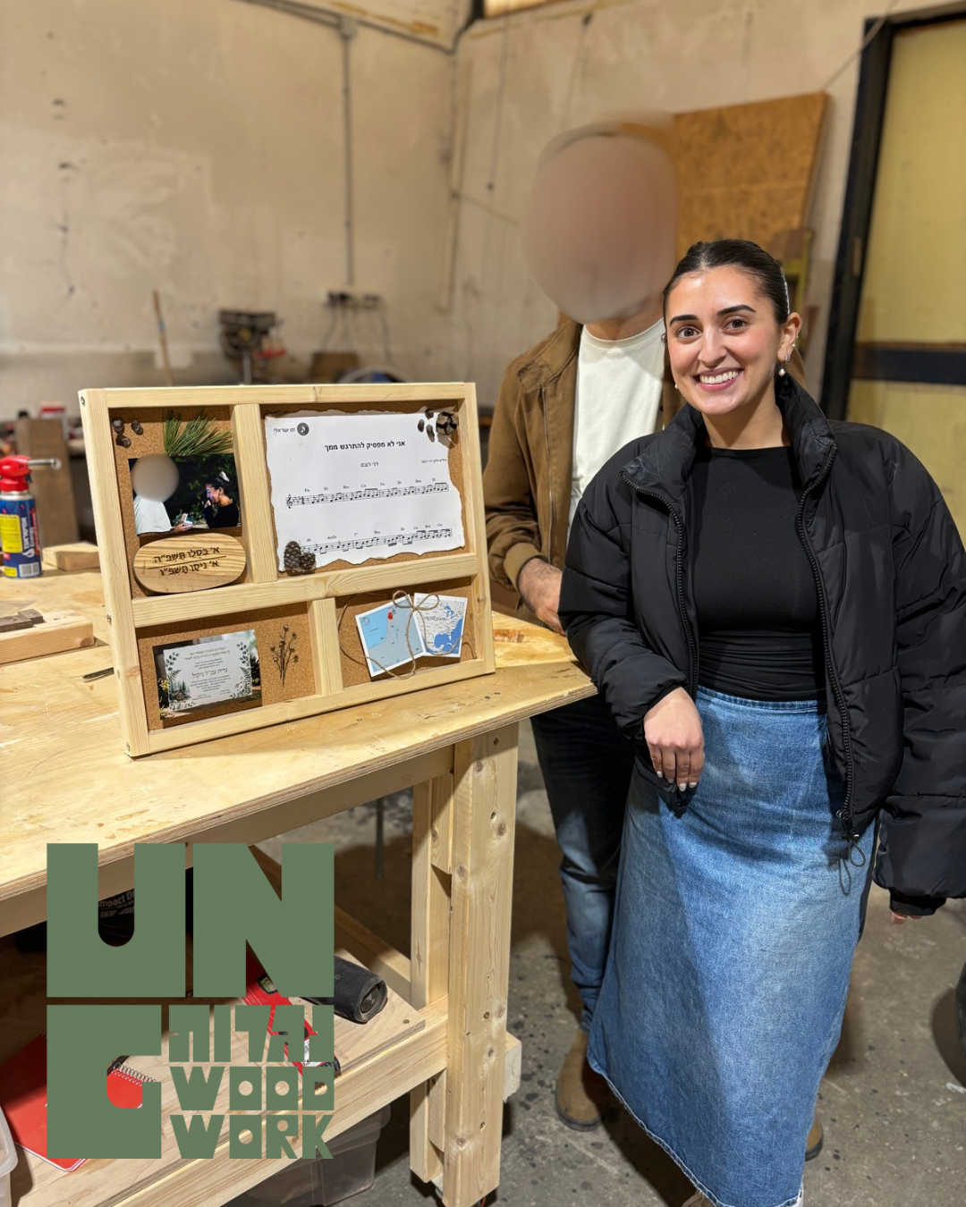 A young woman smiling and posing next to a wooden display with pictures, a note, and decorative items on a workbench in a woodworking workshop.